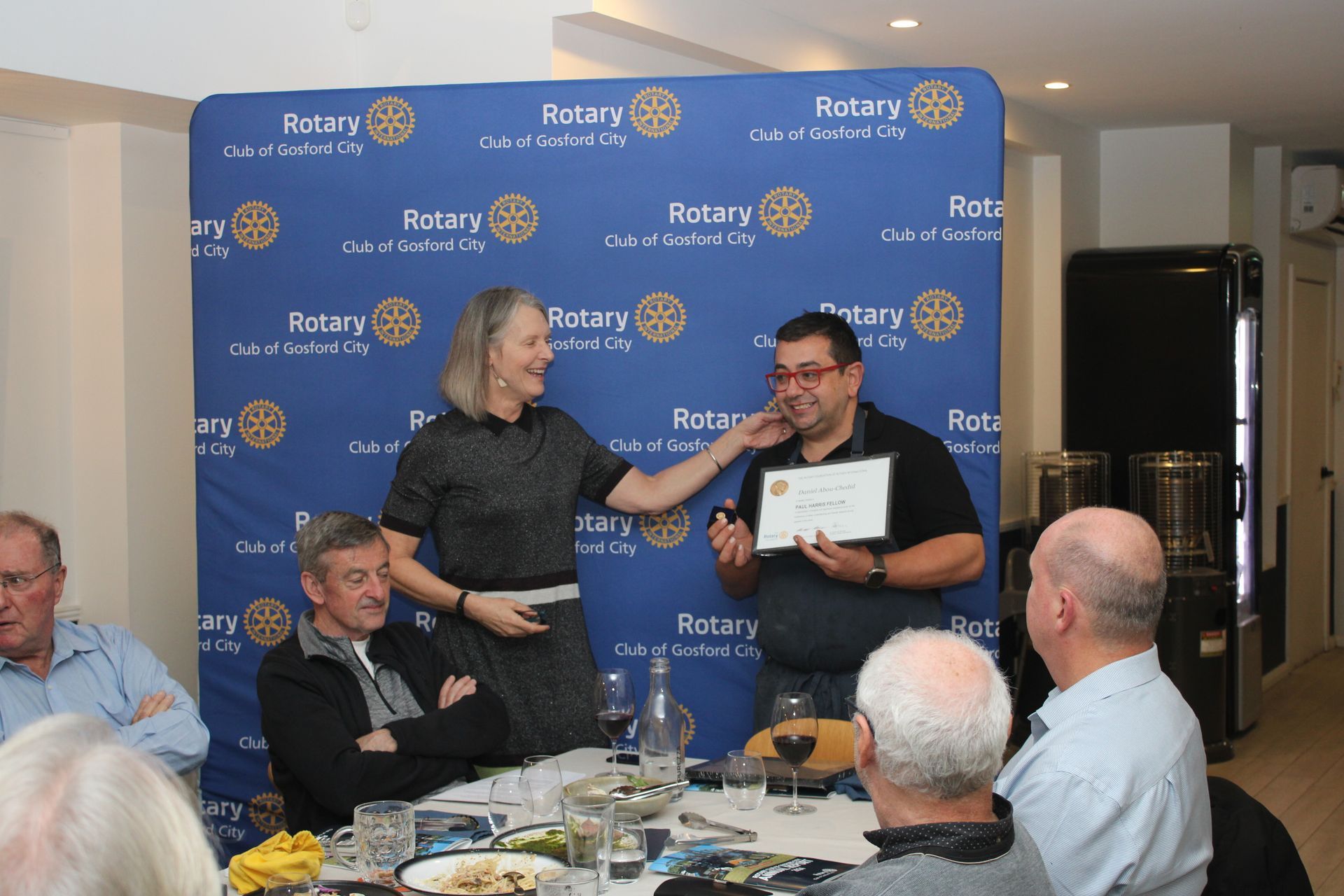 A group of people are sitting around a table in front of a wall that says rotary