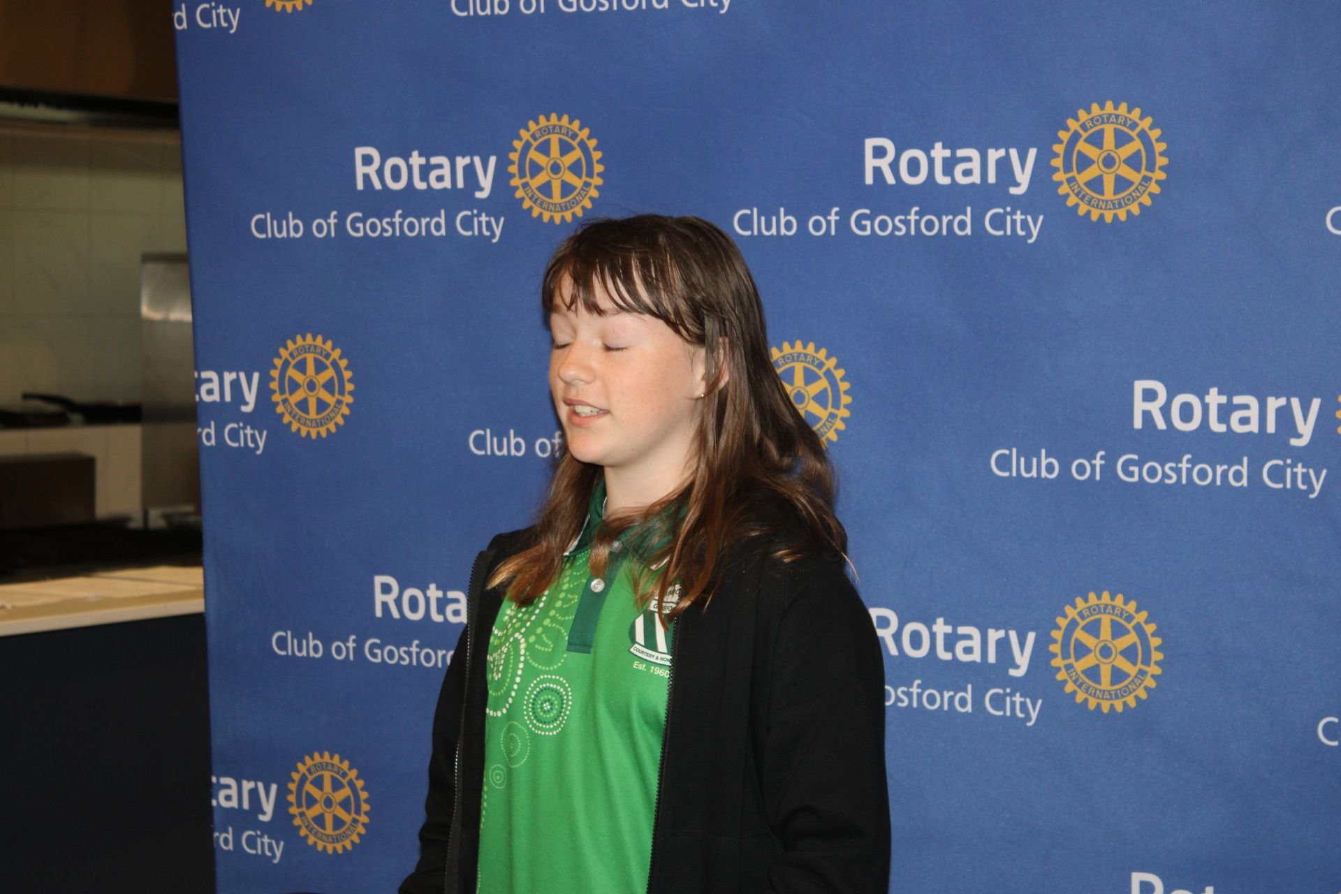 A girl in a green shirt is standing in front of a blue background that says rotary
