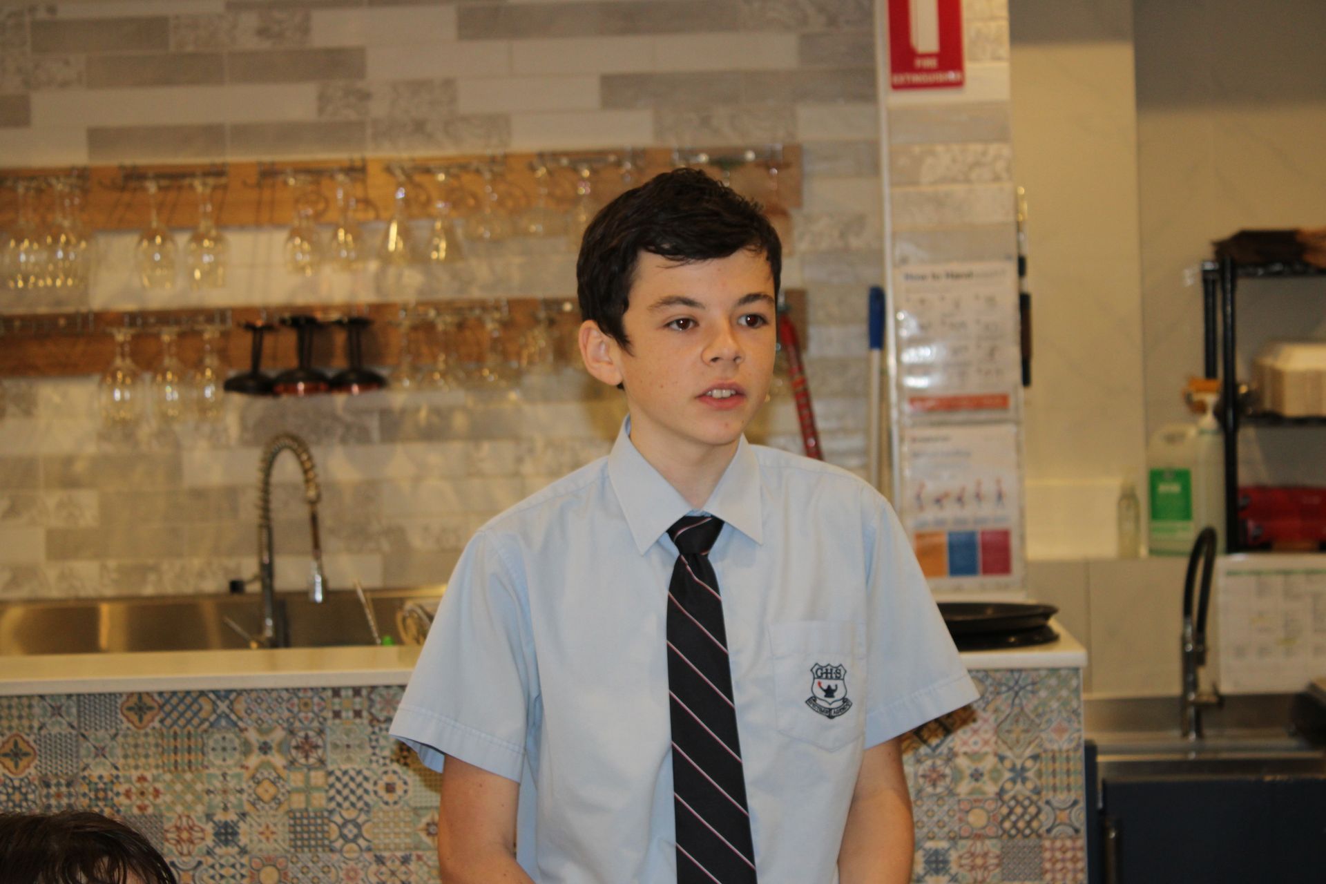 A young boy in a blue shirt and black tie is standing in a kitchen.