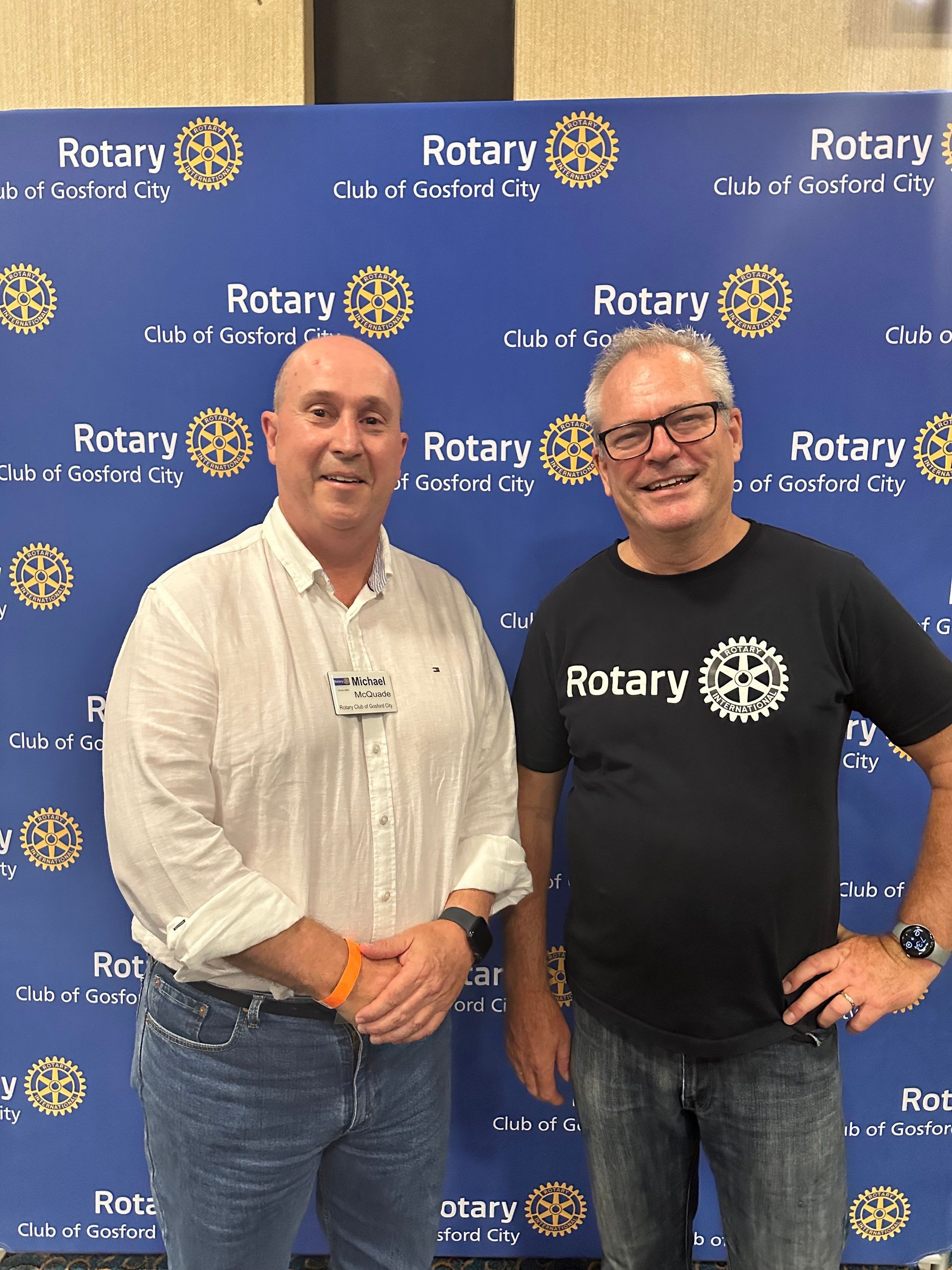 Two men are posing for a picture in front of a wall that says rotary