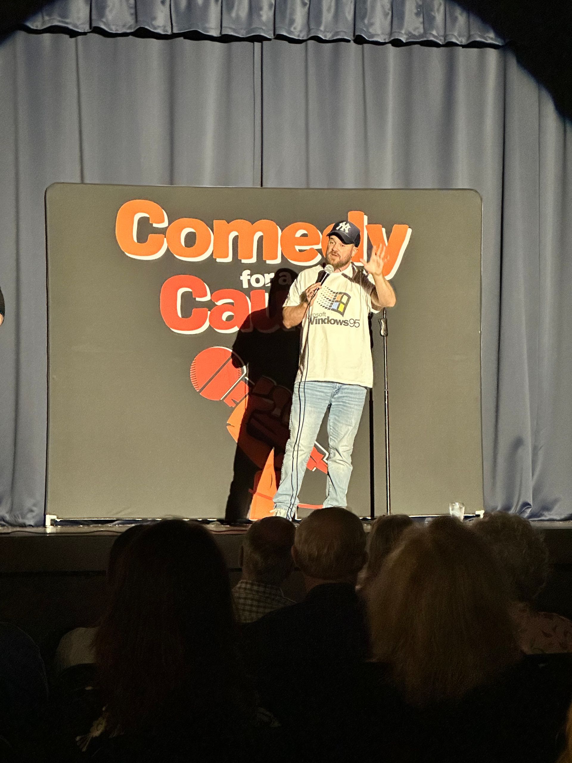 A man stands on a stage in front of a comedy for cancer sign