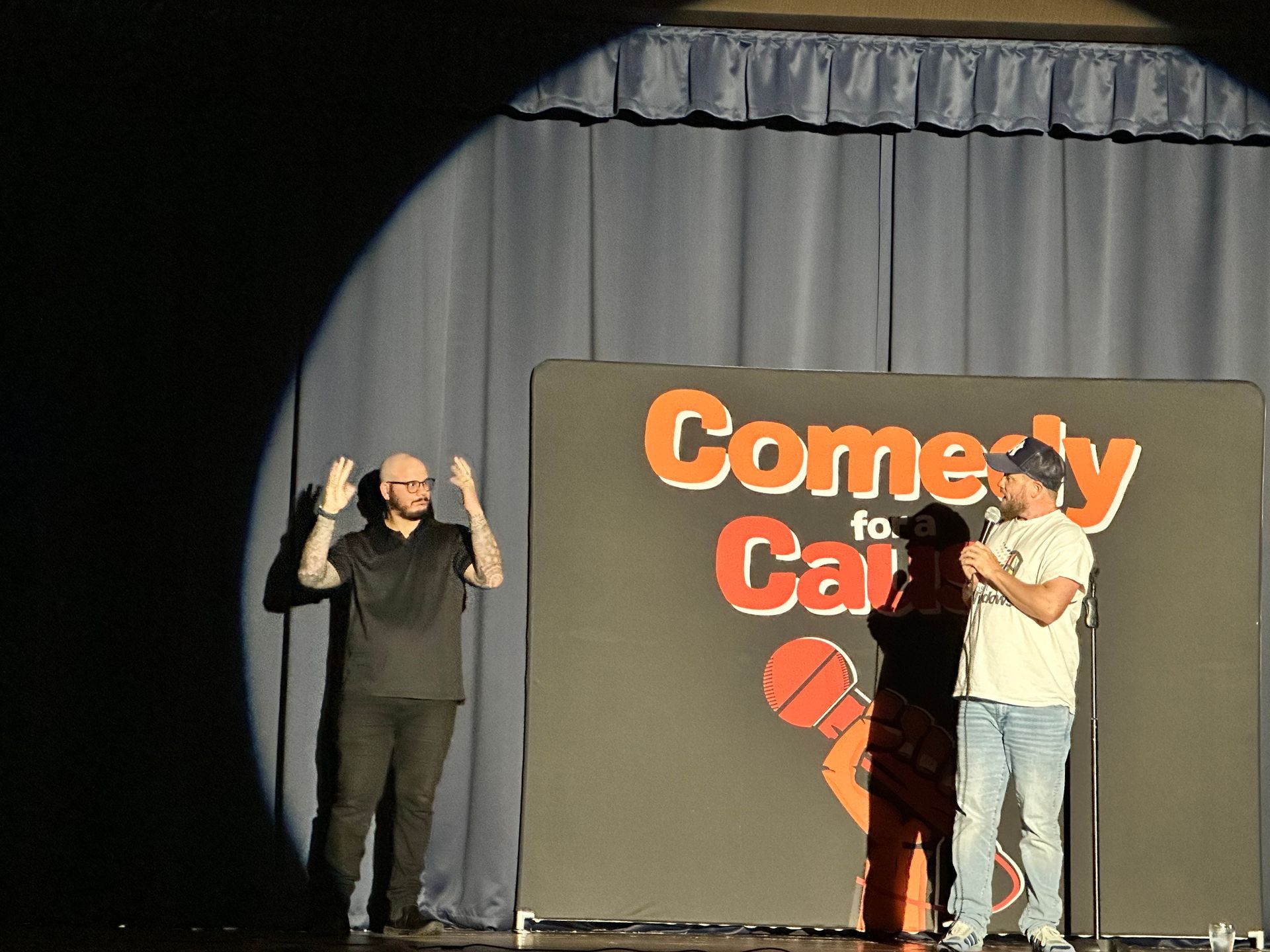 Two men stand on a stage in front of a sign that says comedy for cars