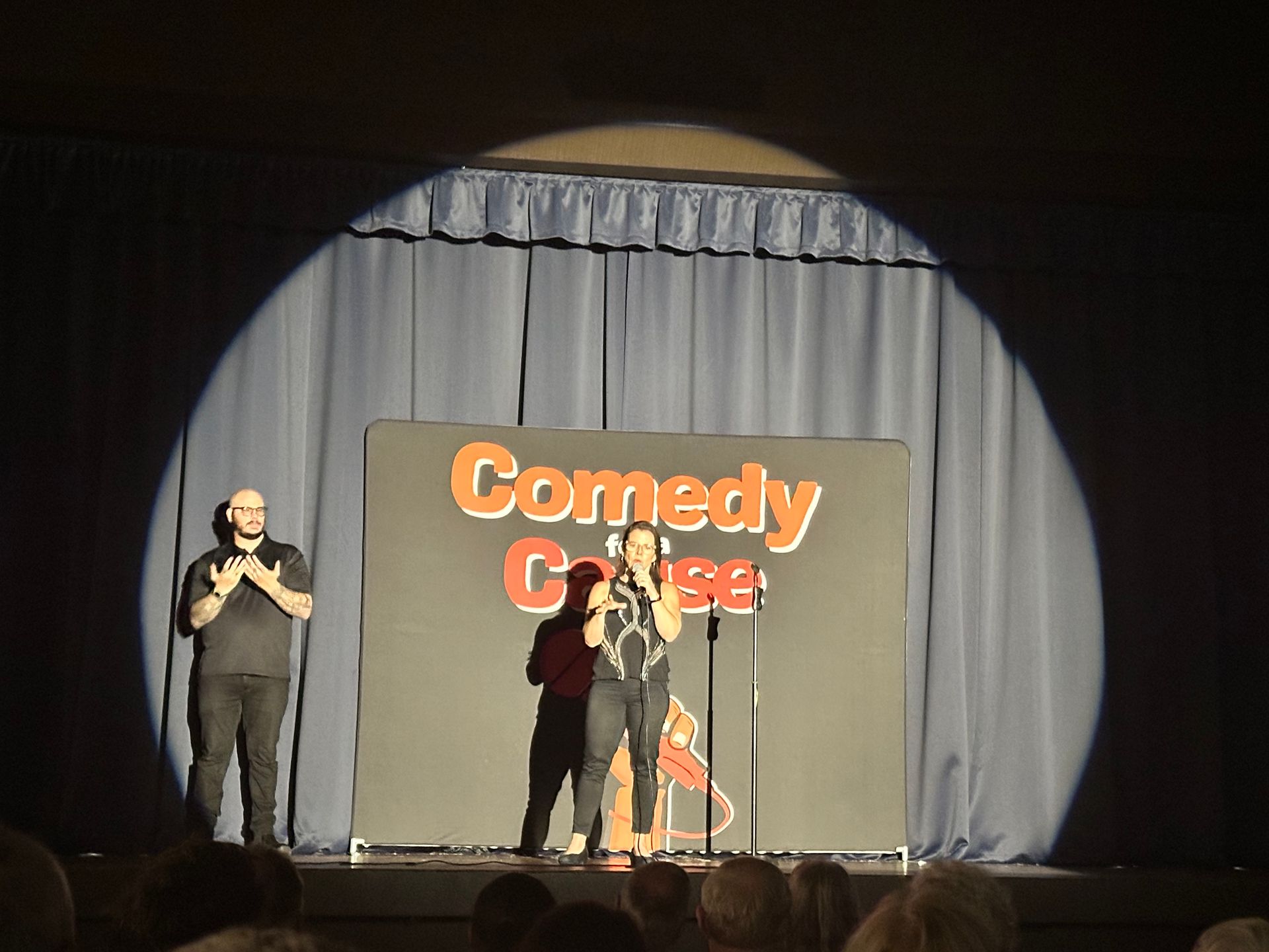 Two people stand on a stage in front of a sign that says comedy classes