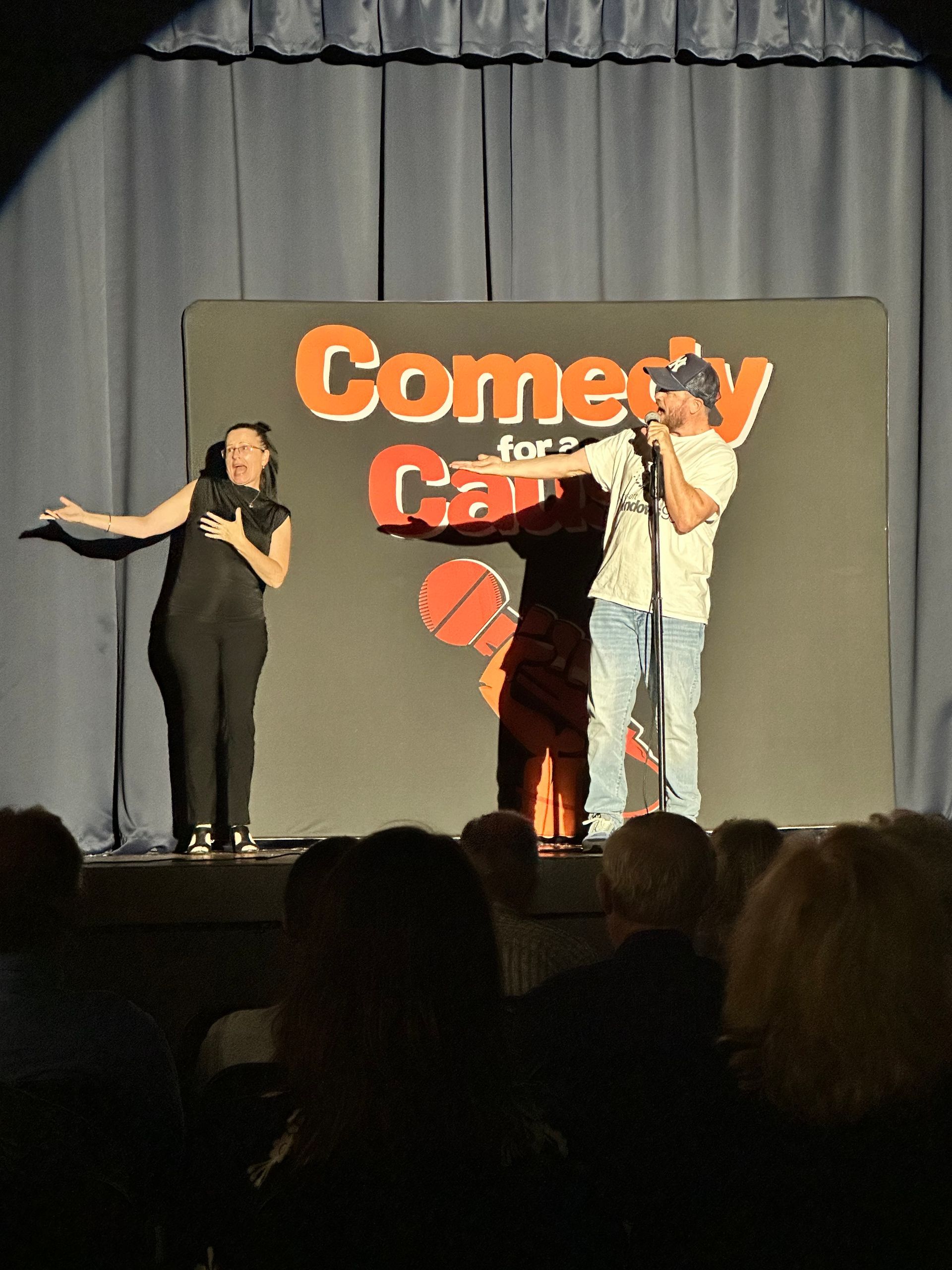 A man and a woman stand on a stage in front of a sign that says comedy for cancer