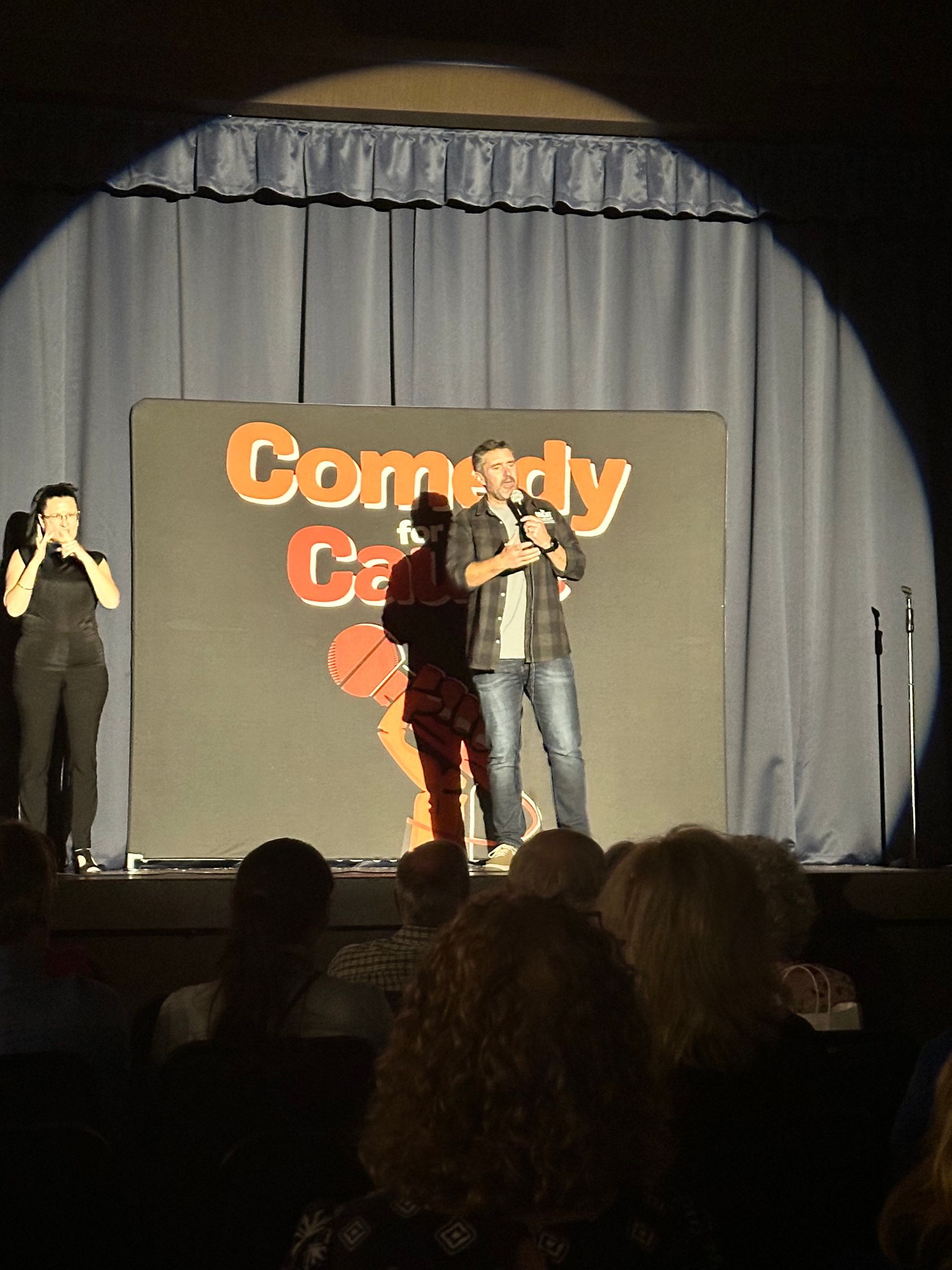 A man and woman stand on a stage in front of a sign that says comedy camp
