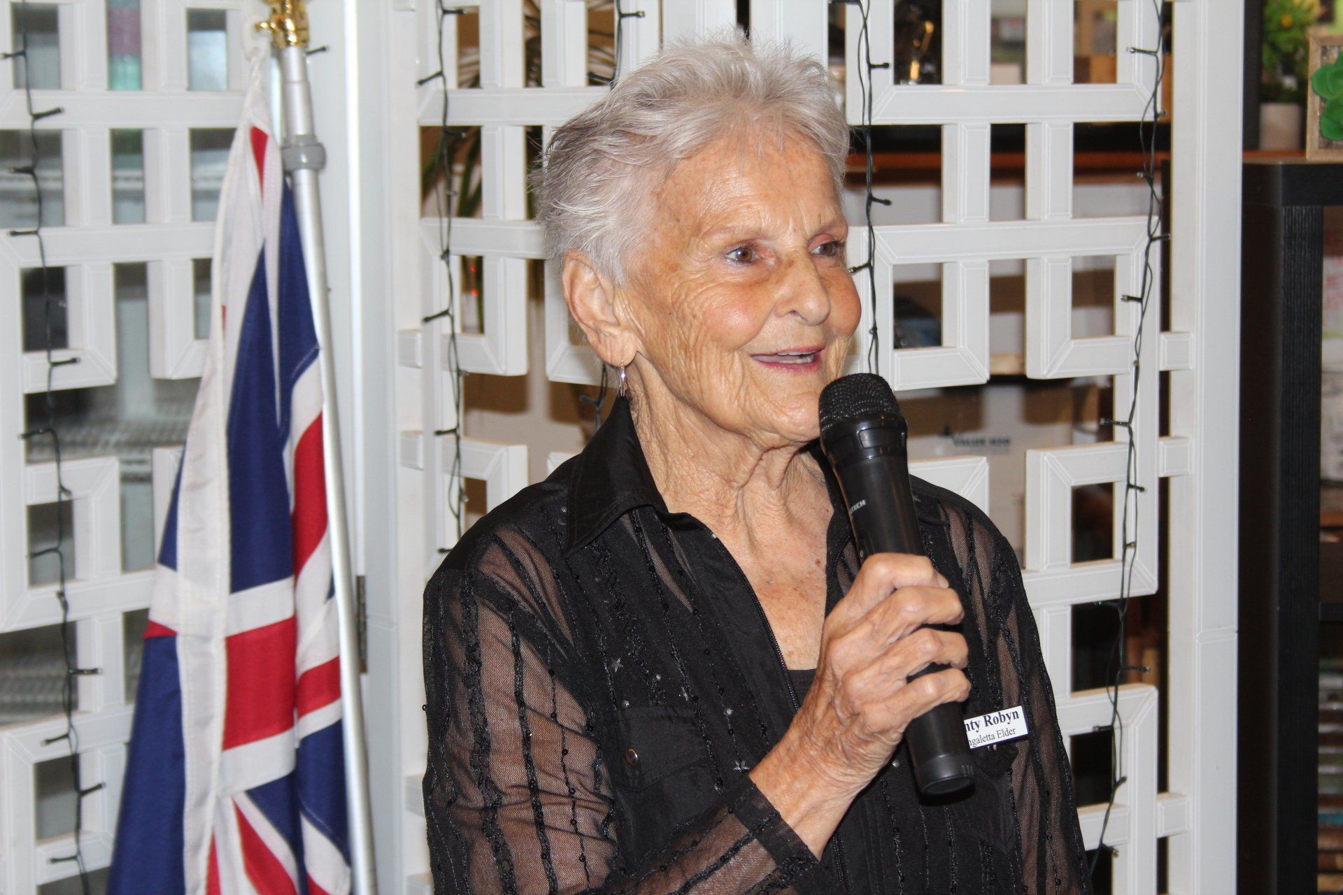 An elderly woman is holding a microphone in front of a british flag.
