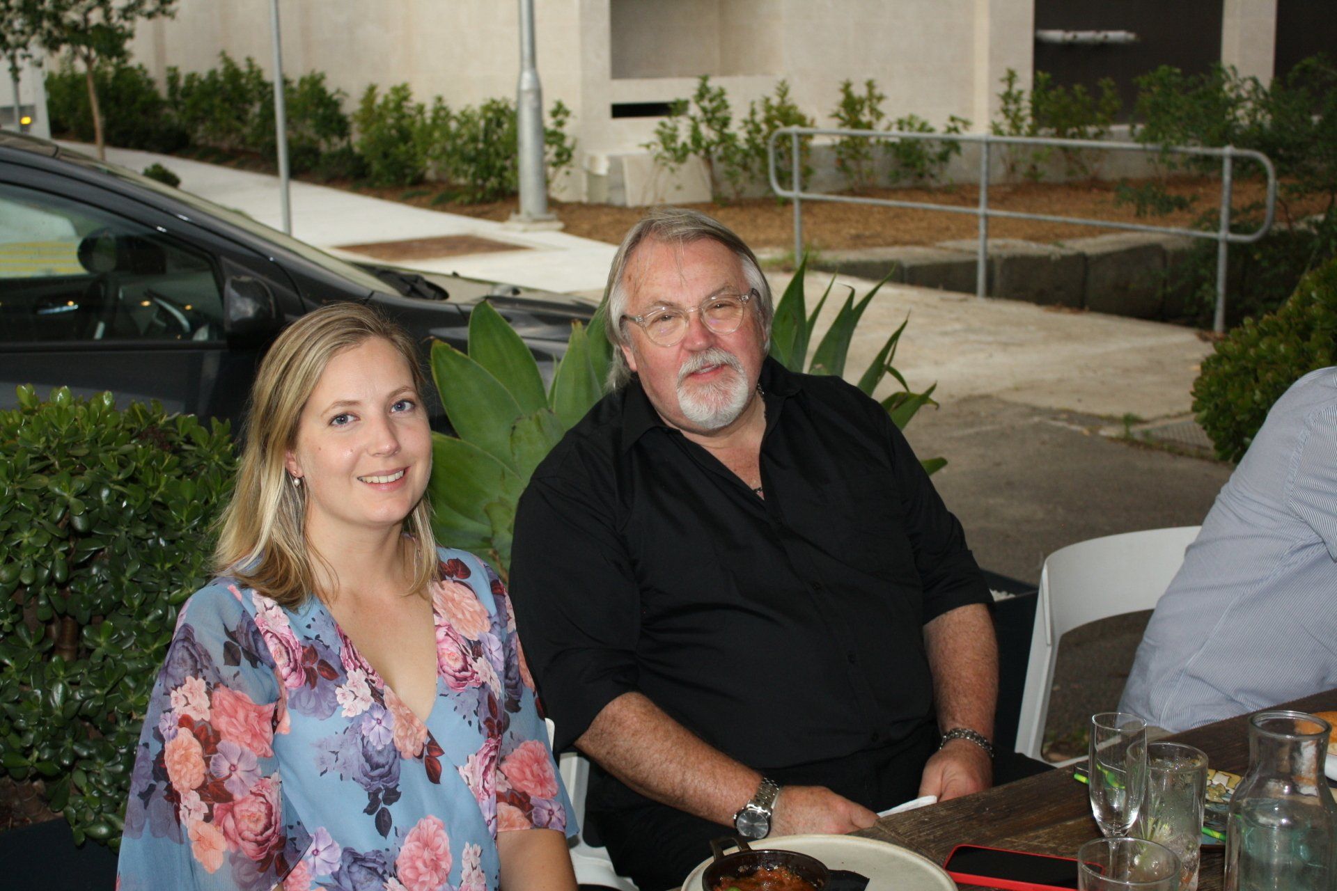 A man and a woman are sitting at a table in front of a car.