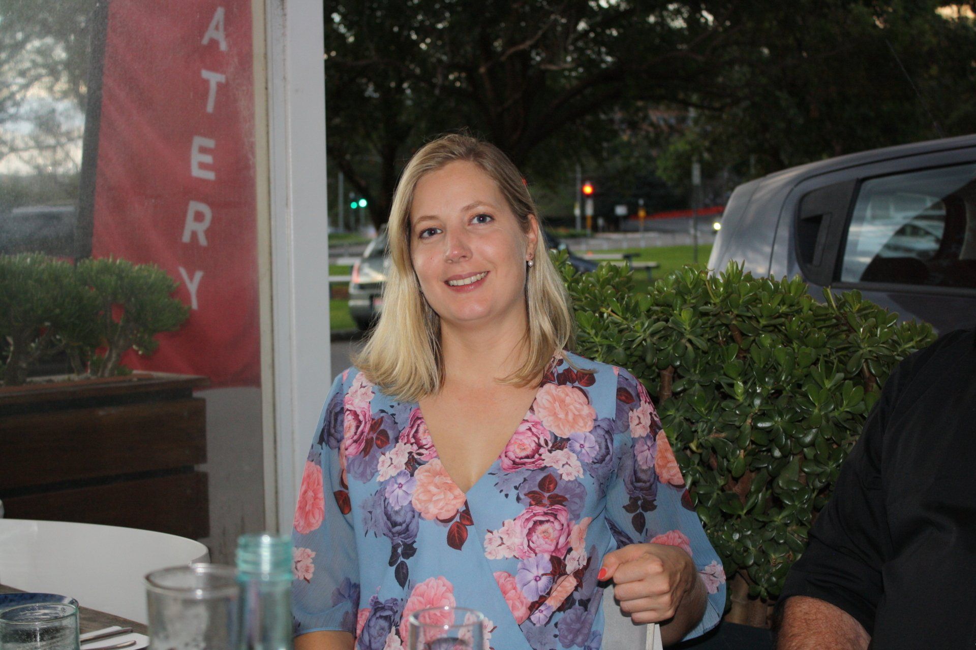 A woman in a floral shirt is sitting at a table with a glass of water.