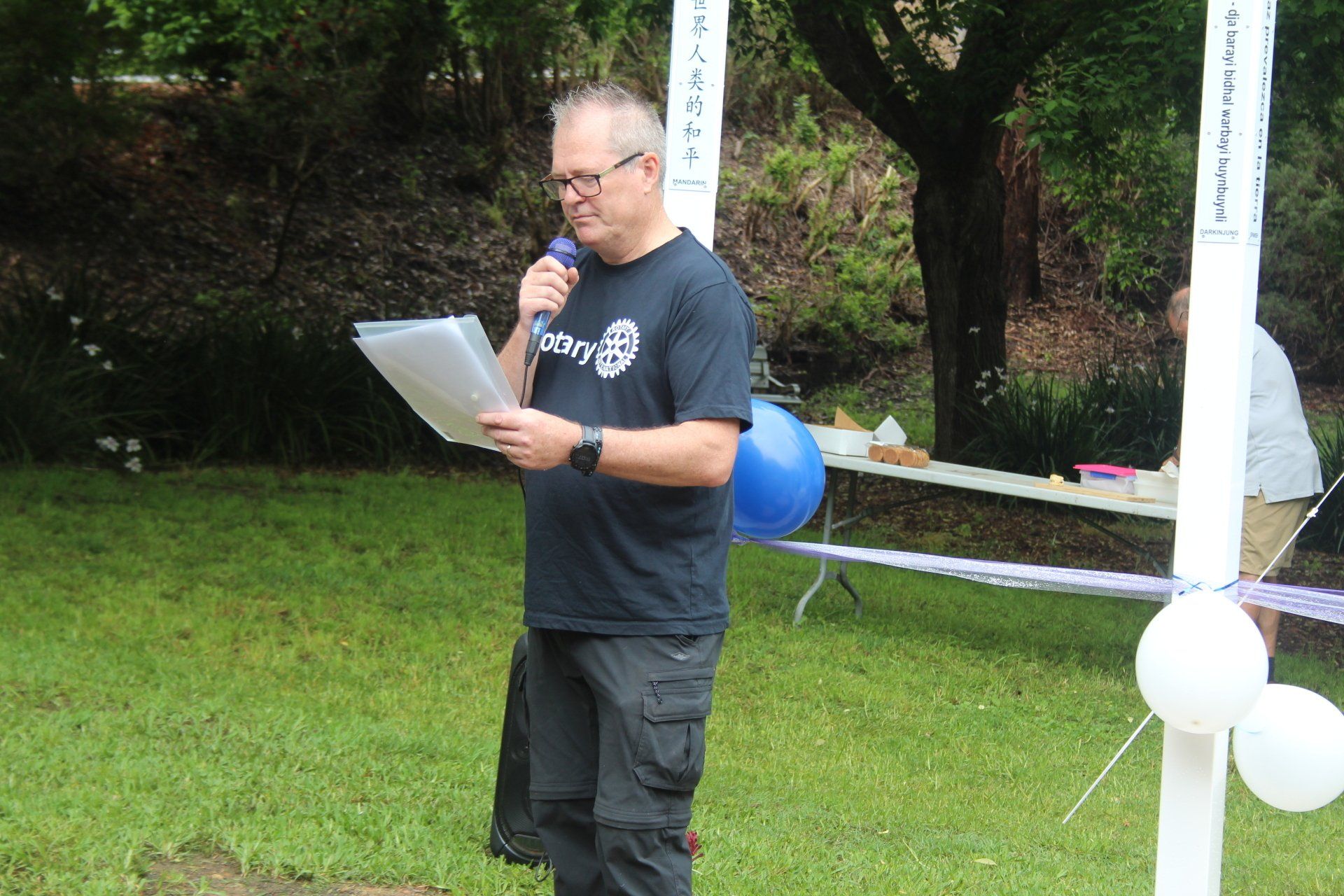 A man is standing in the grass holding a microphone and a piece of paper.