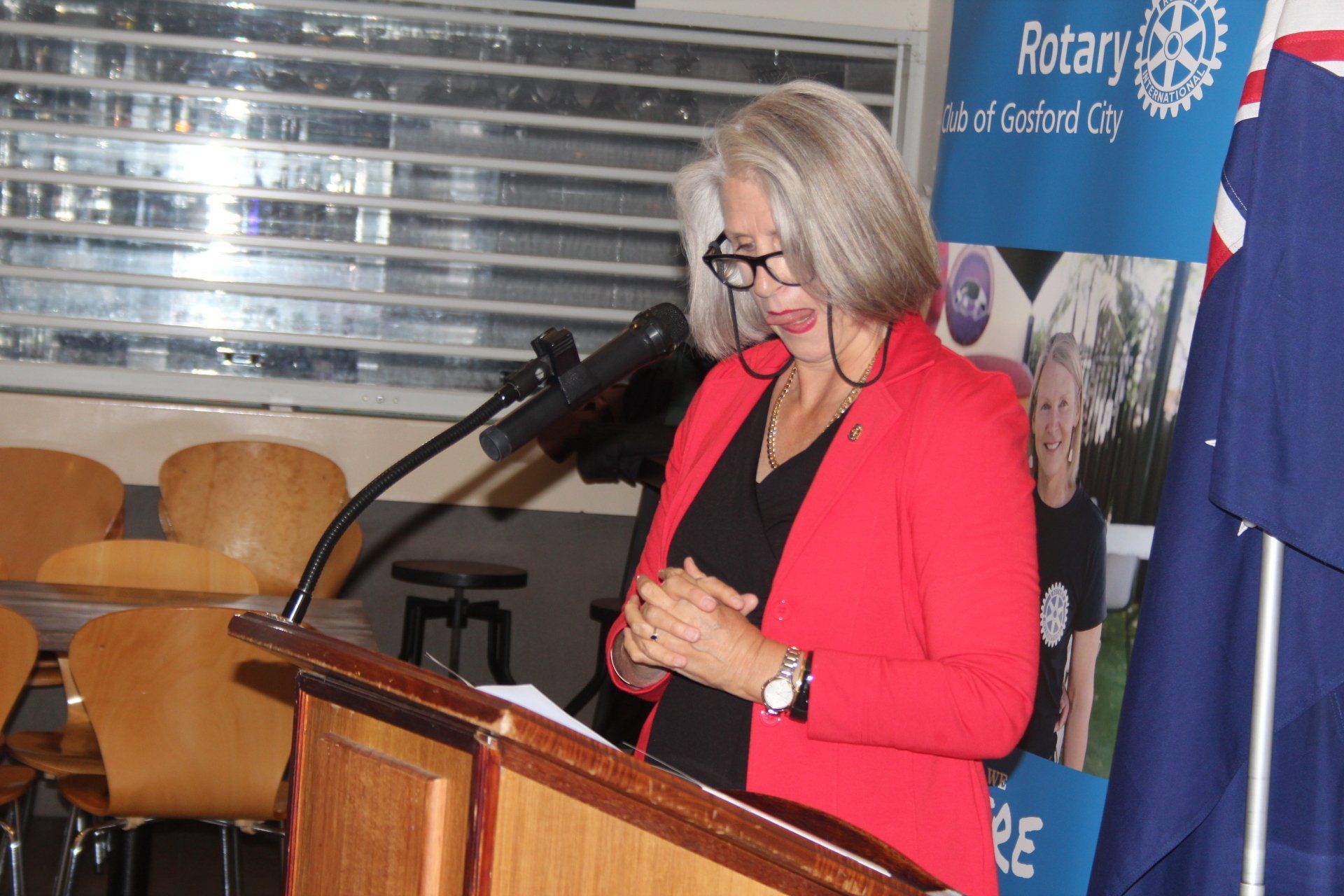 A woman in a red jacket is standing at a podium speaking into a microphone.