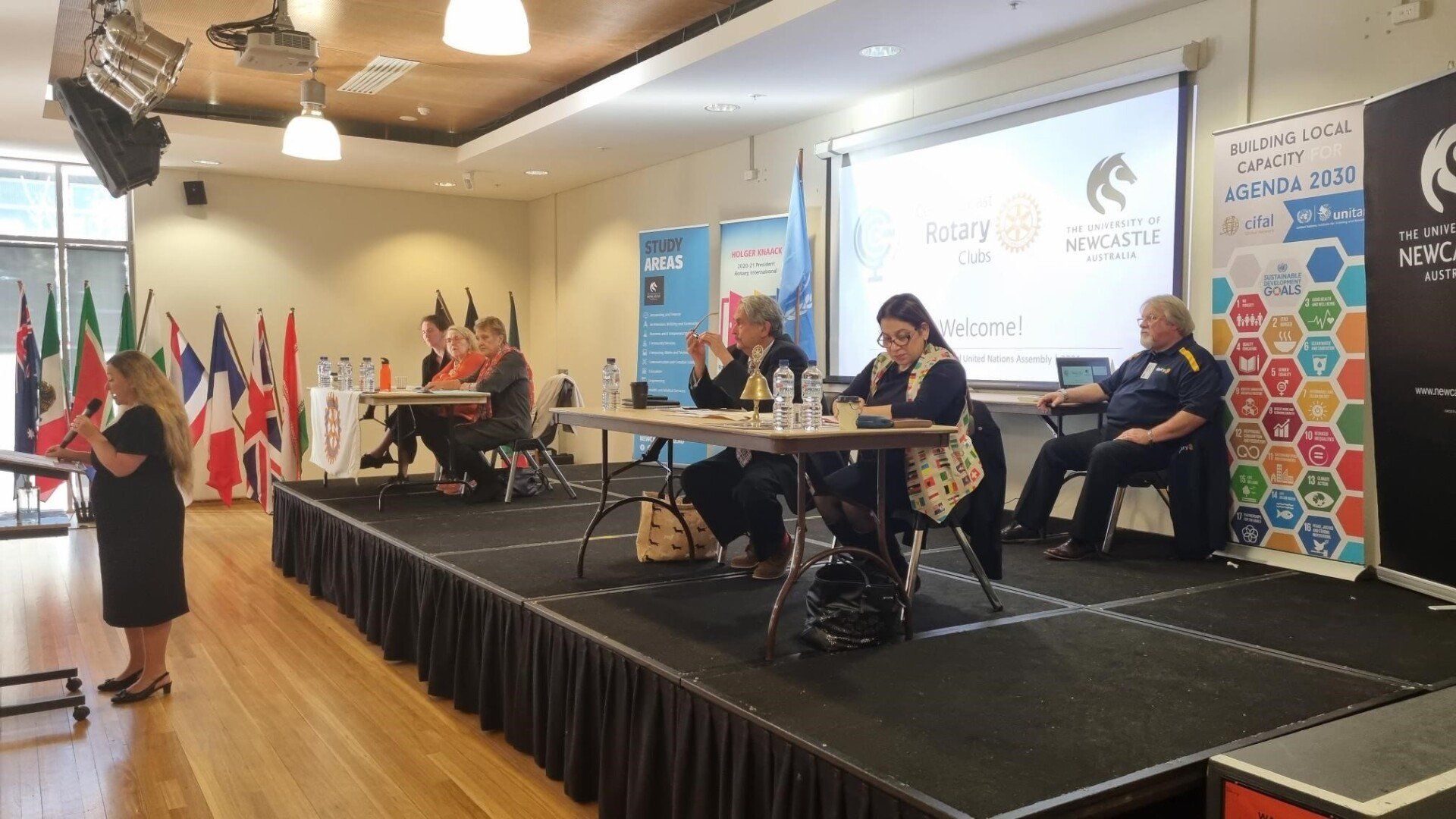 A woman is standing on a stage in front of a group of people sitting at tables.