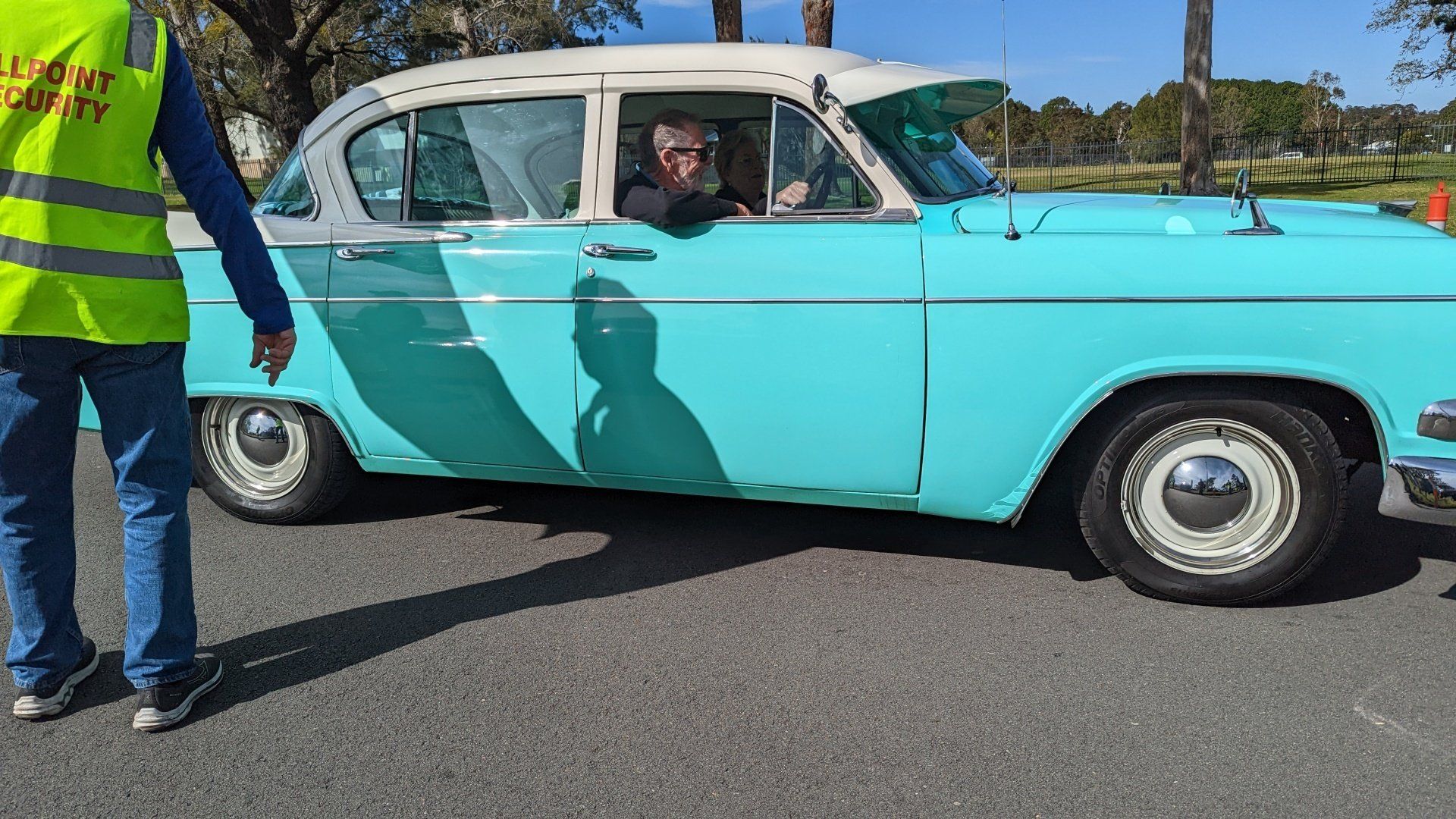 A man in a yellow vest is standing next to a blue and white car.