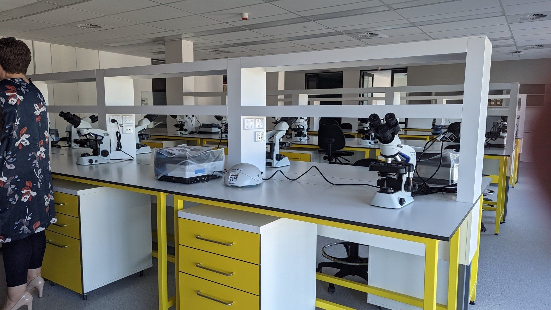 A woman is standing in front of a row of microscopes in a lab.