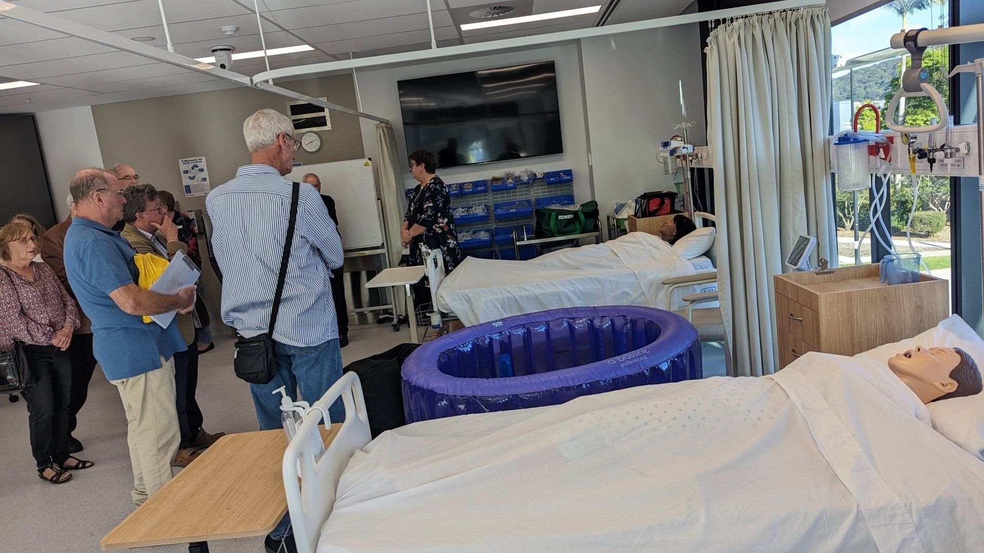A group of people are standing around a hospital room with a mannequin in a hospital bed.