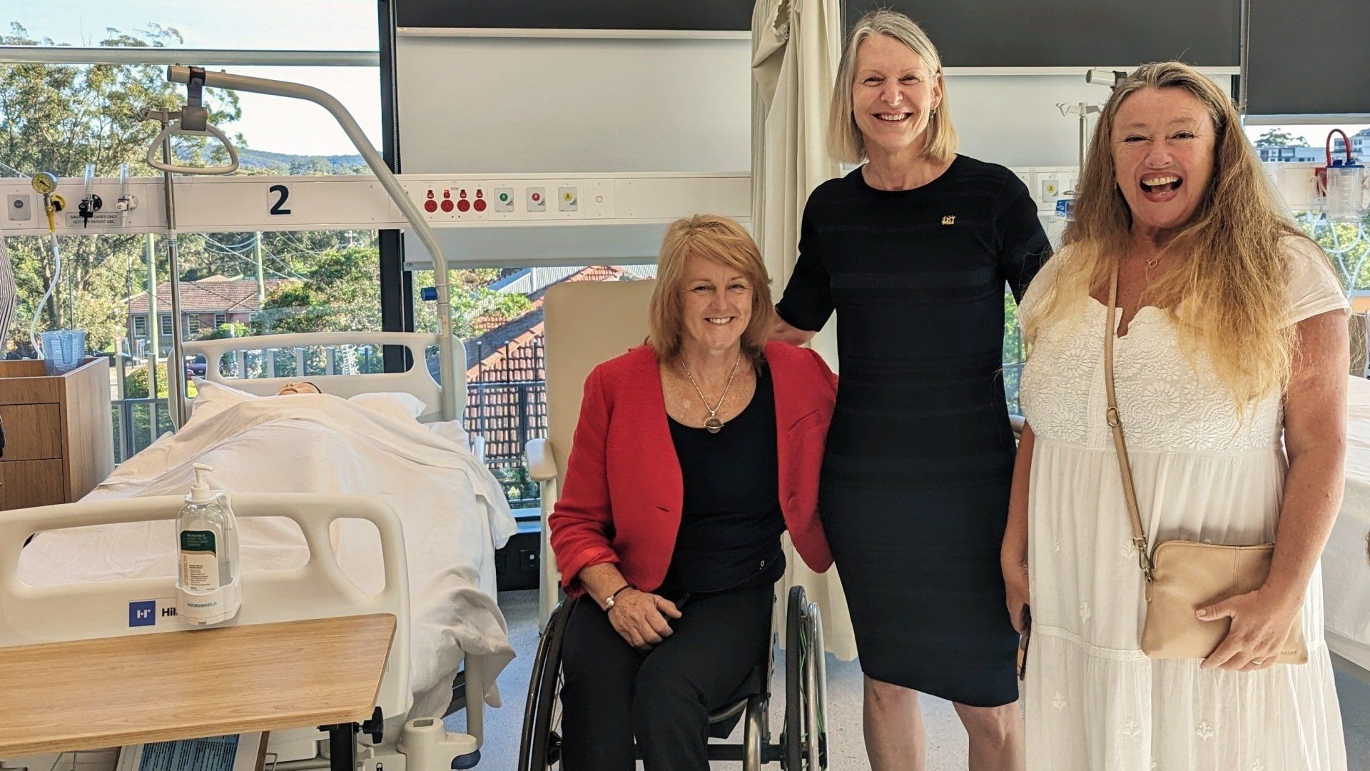 Three women are standing next to a woman in a wheelchair in a hospital room.