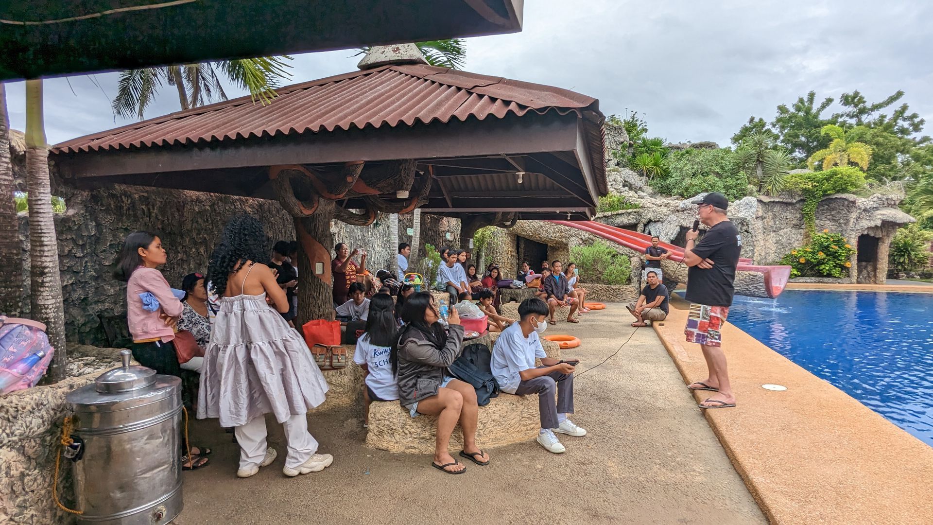A group of people are sitting under a pavilion next to a swimming pool.