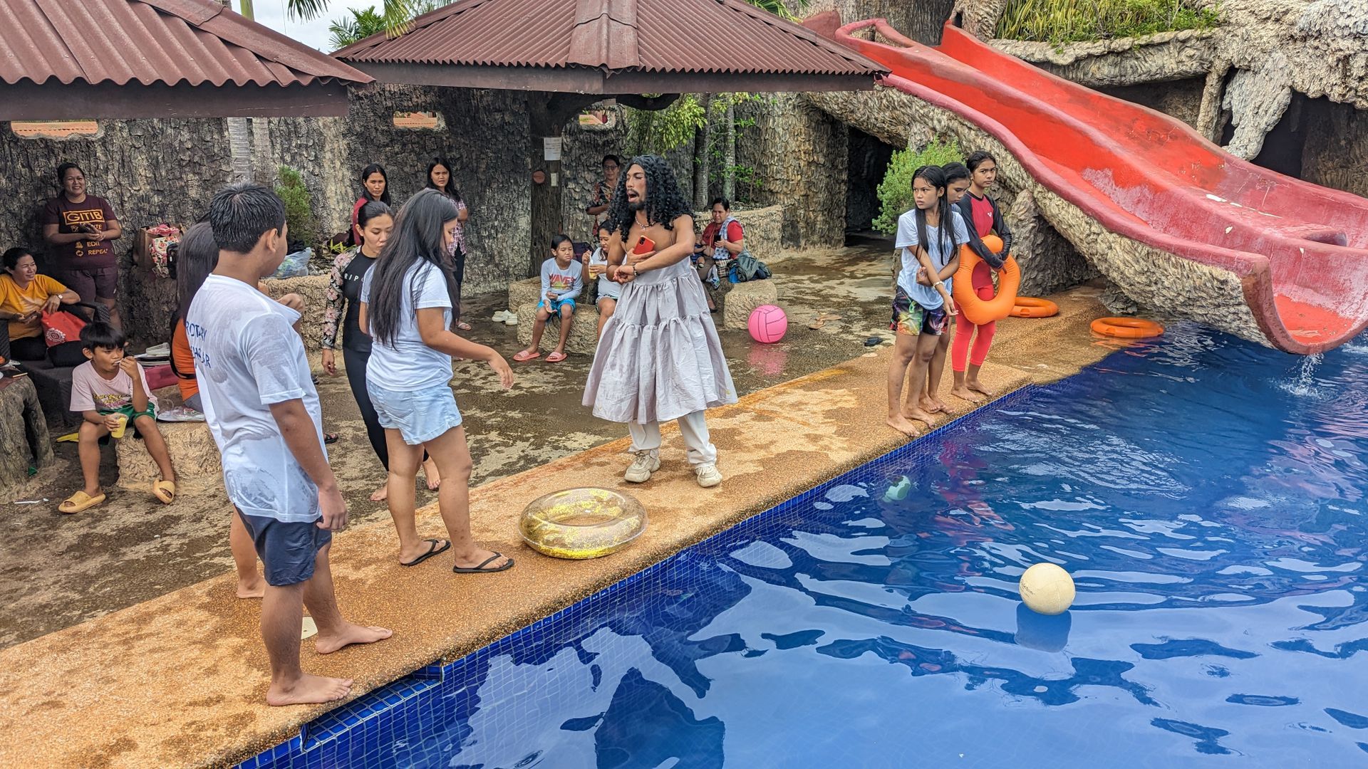 A group of people are standing around a swimming pool with a red slide.