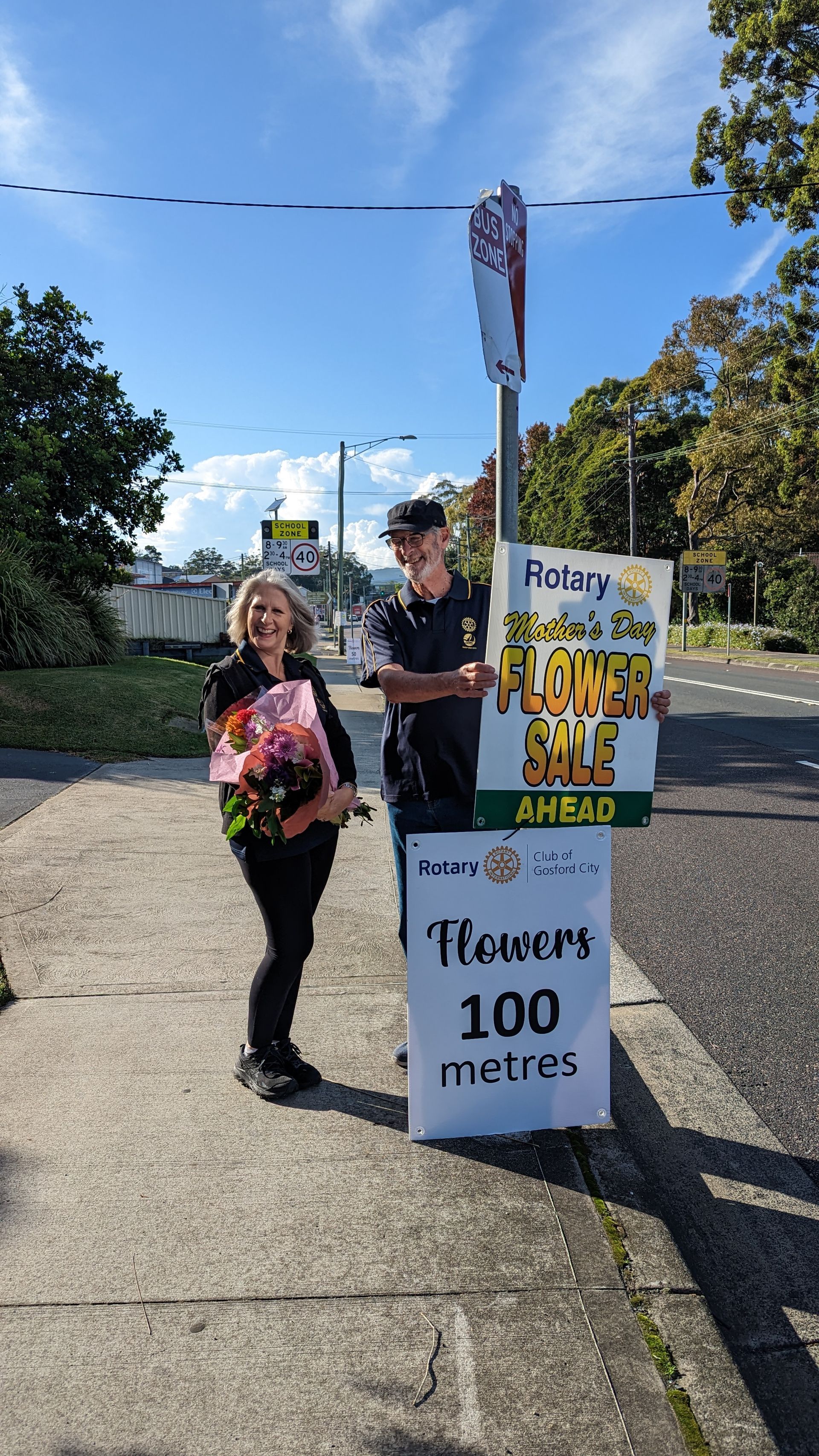 A man and a woman are standing next to a sign that says `` flowers 100 metres ''.
