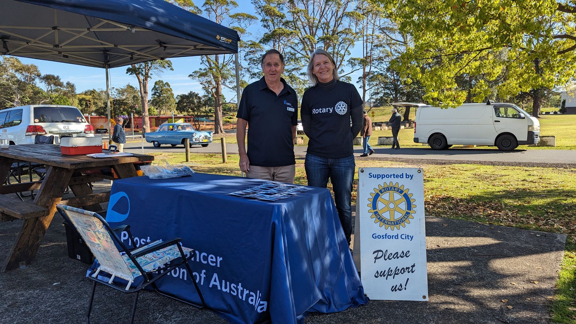 Two men are standing in front of a table in a park.