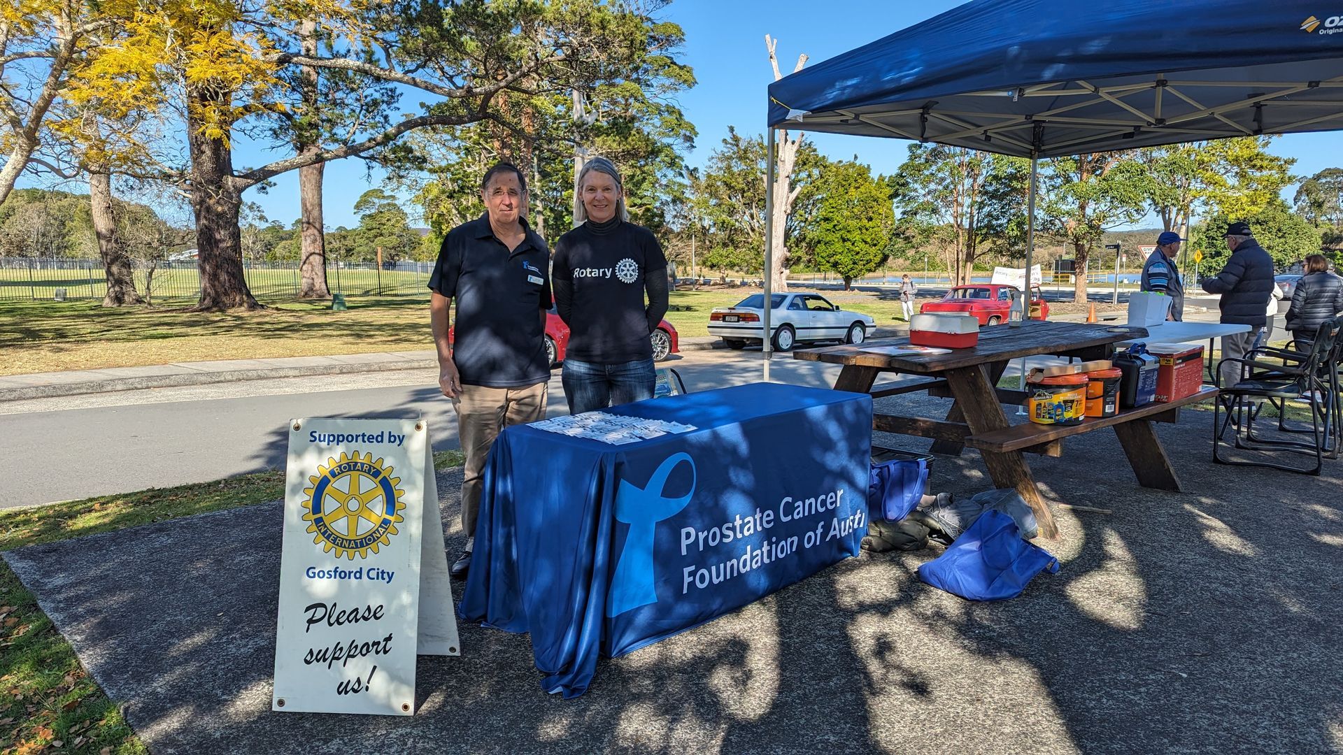 Two men are standing in front of a table in a park.