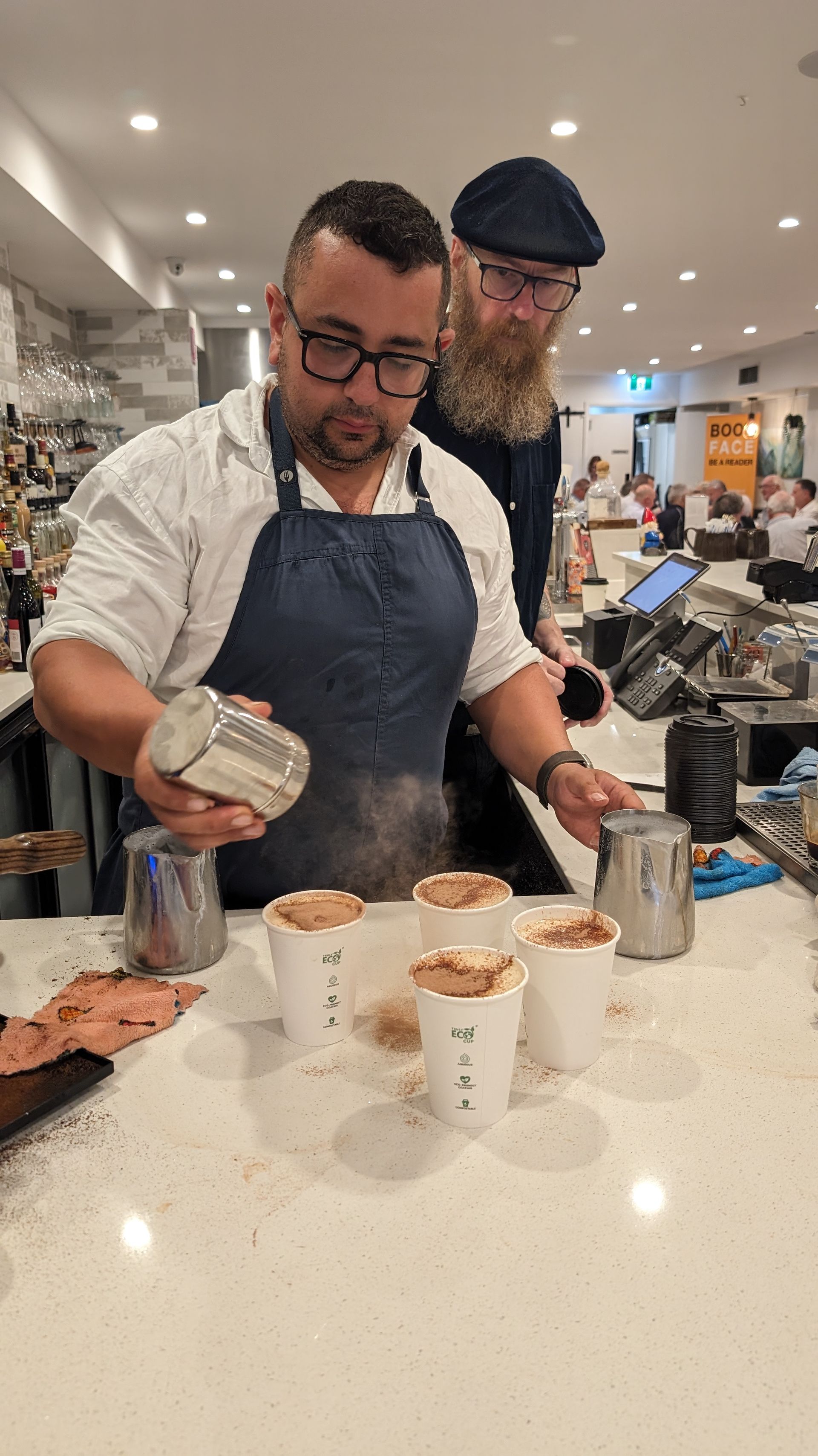 A man is pouring milk into a cup of coffee while another man watches.