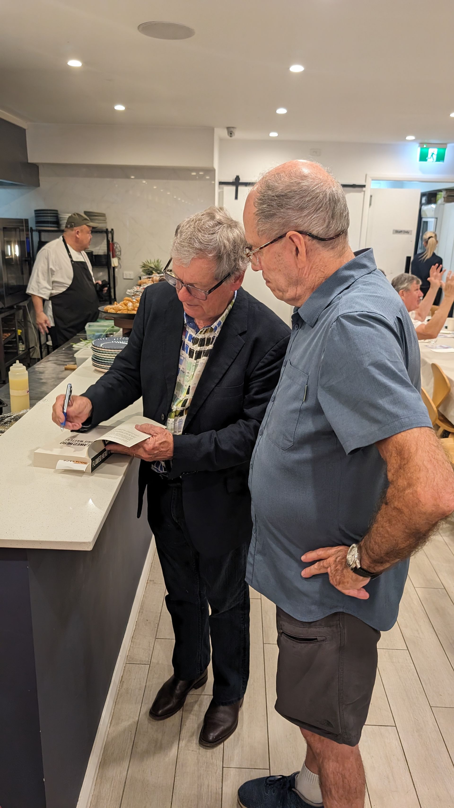 Two men are standing next to each other in a kitchen signing a book.