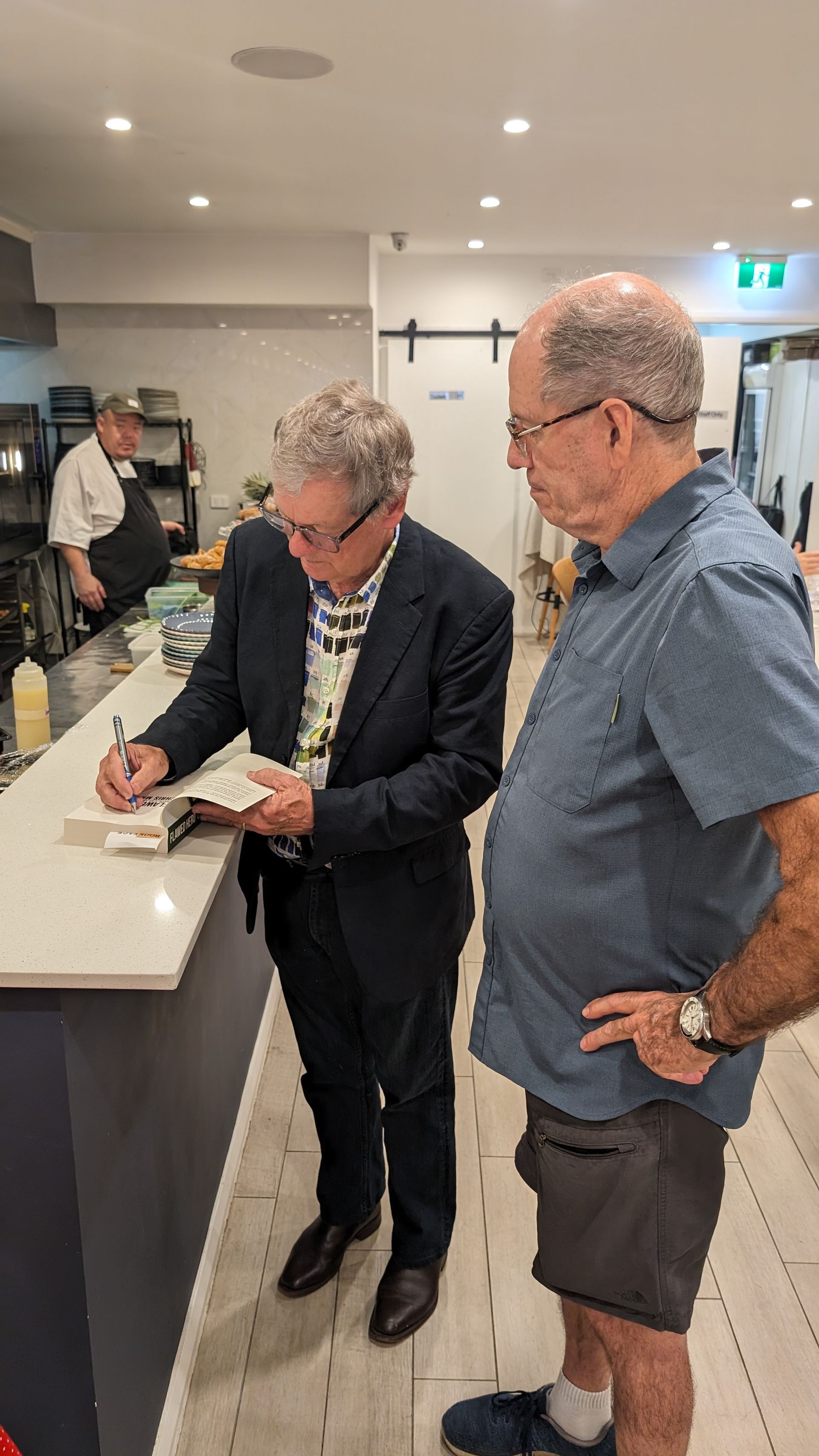 Two men are standing next to each other in a kitchen . one of the men is signing a book.