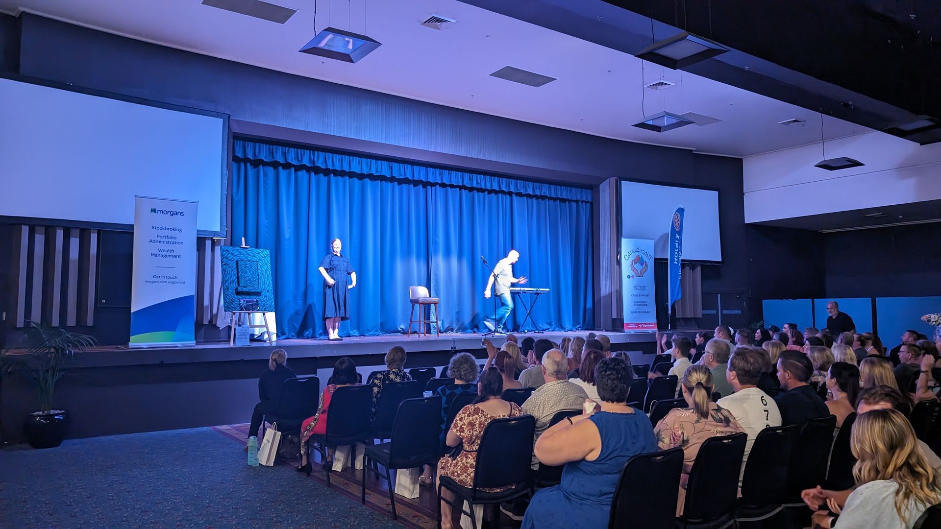 A group of people are sitting in front of a stage with a blue curtain.
