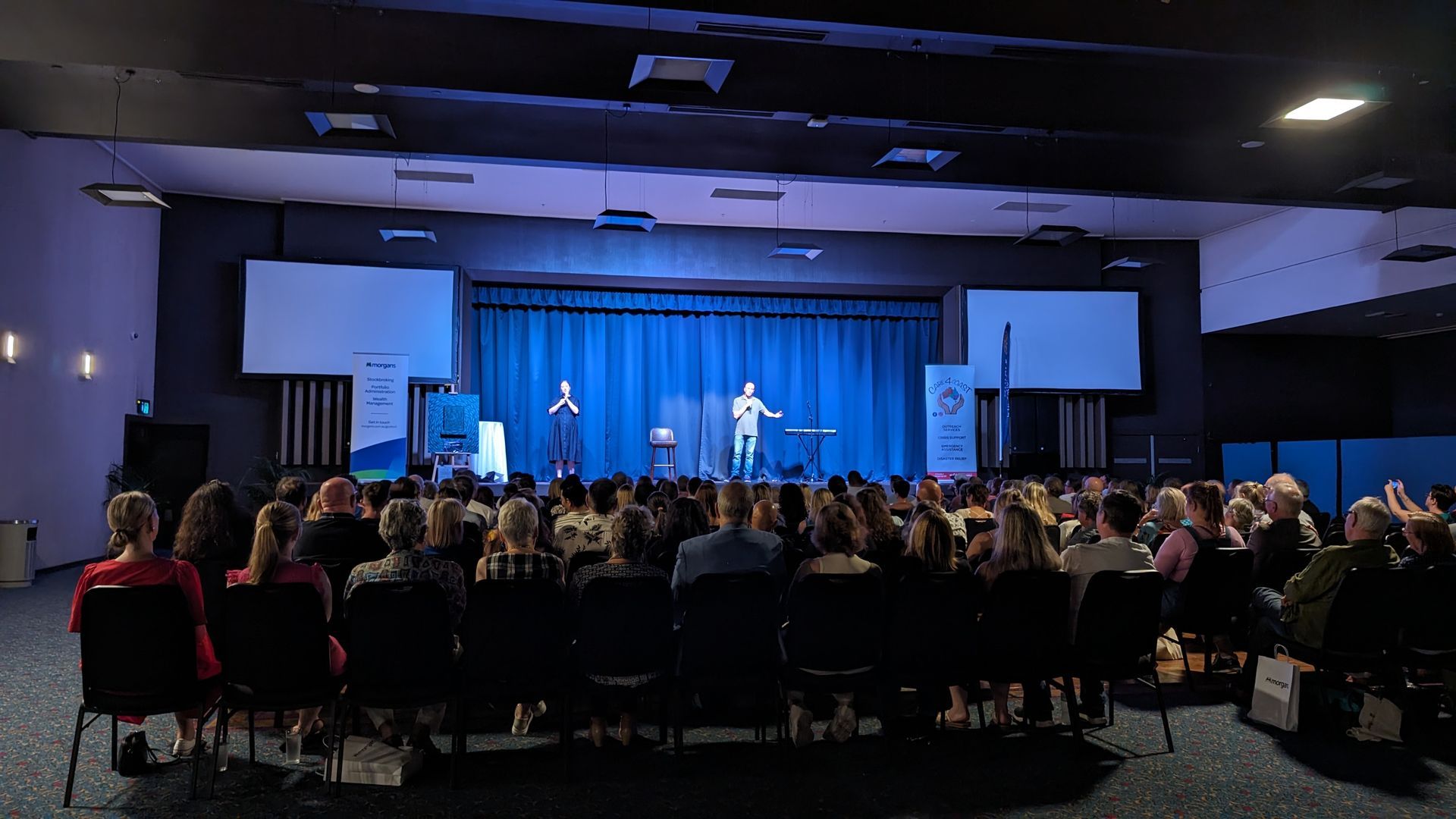 A large auditorium with a blue curtain and people sitting in chairs.