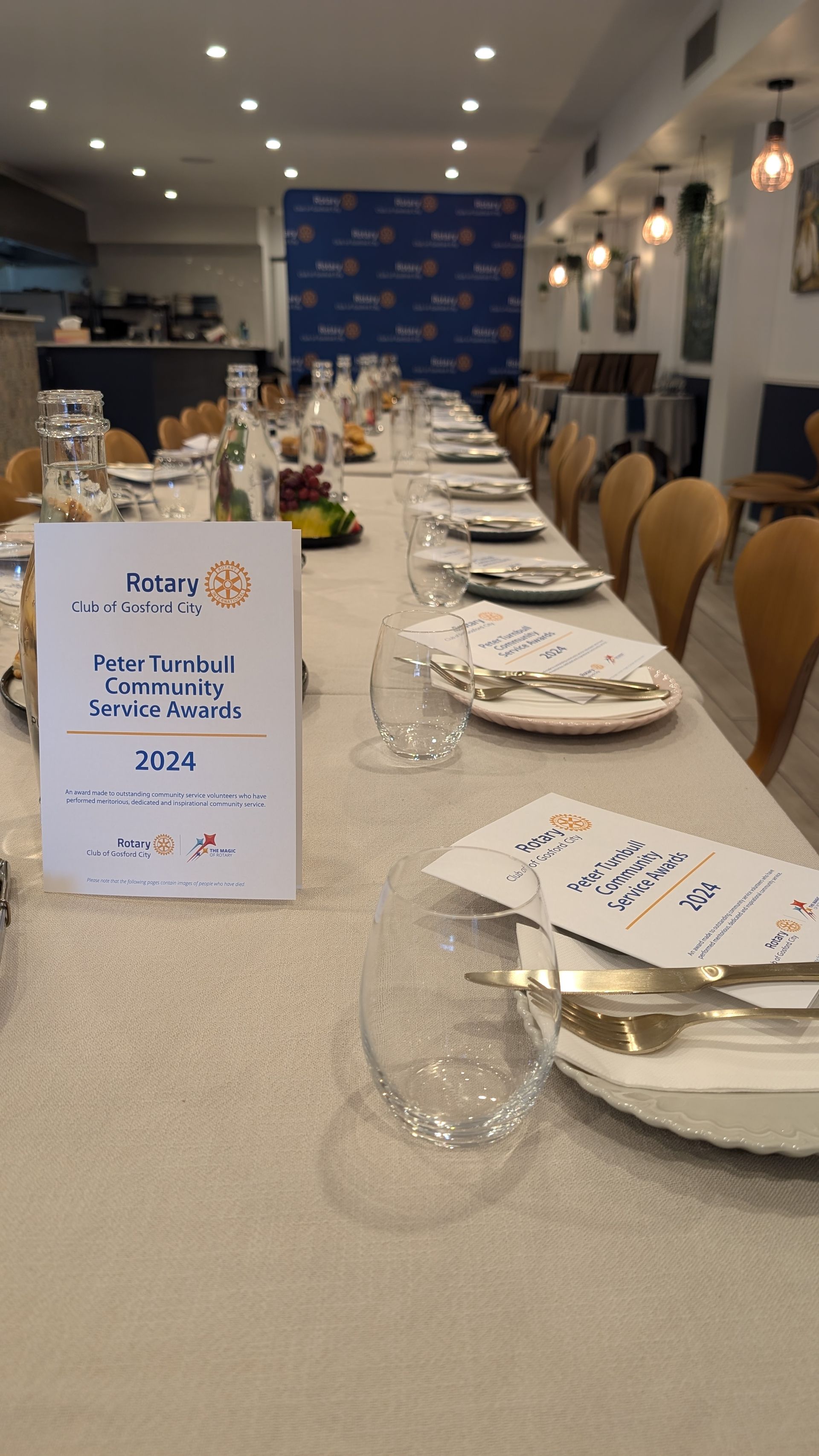 A long table with plates , glasses , napkins and a sign on it.