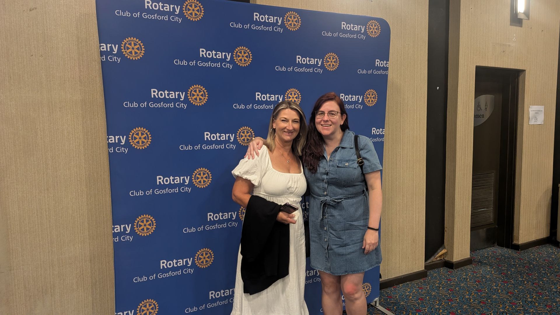 Two women are posing for a picture in front of a blue wall that says rotary.