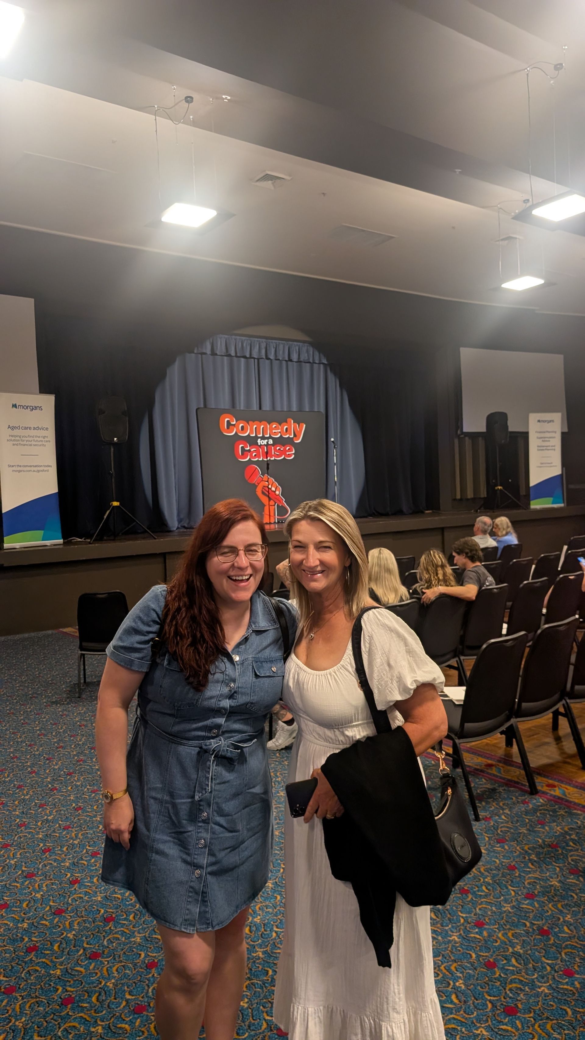 Two women are posing for a picture in front of a stage.