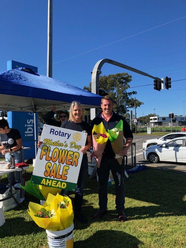 Two people are standing in front of a tent holding a sign that says flower sale ahead.