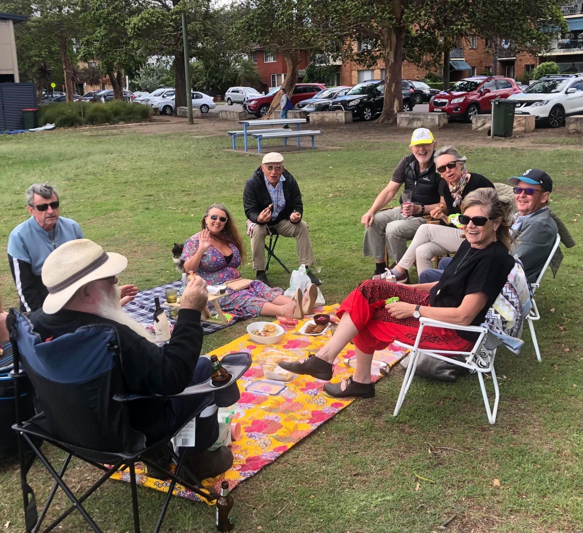 A group of people are sitting around a picnic blanket in a park.