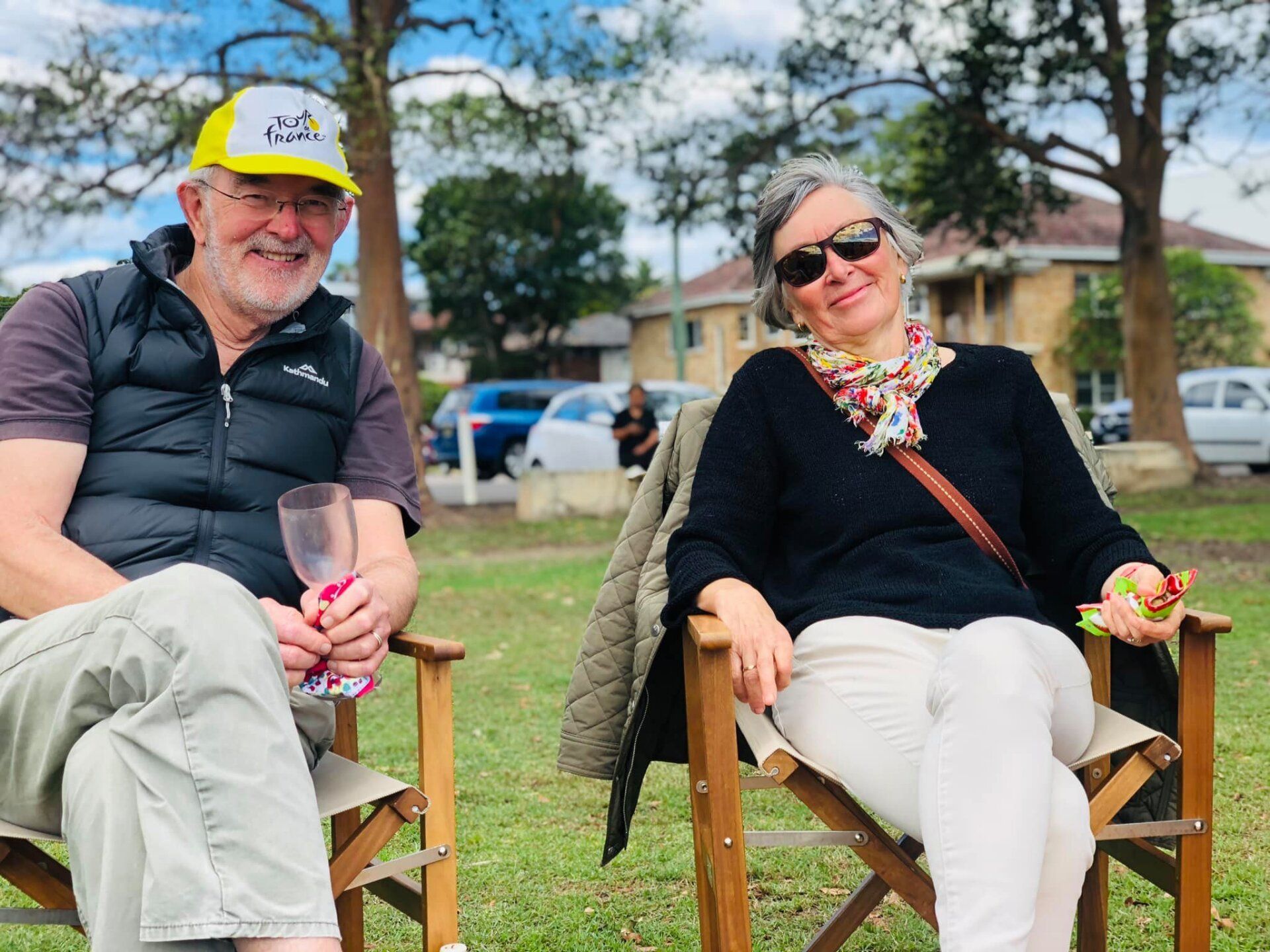 A man and a woman are sitting in chairs in a park holding wine glasses.