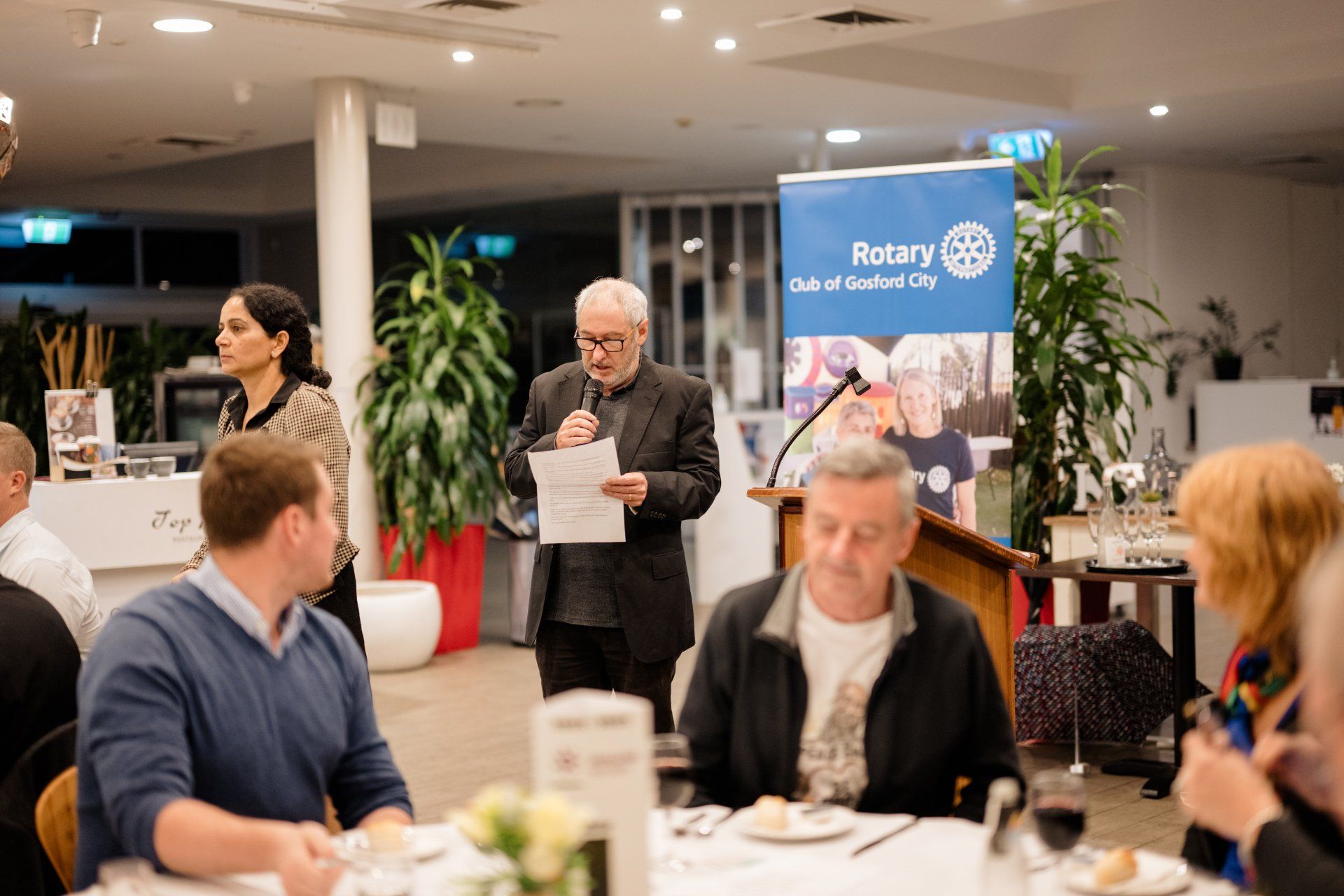 A man is standing at a podium giving a speech to a group of people sitting at a table.