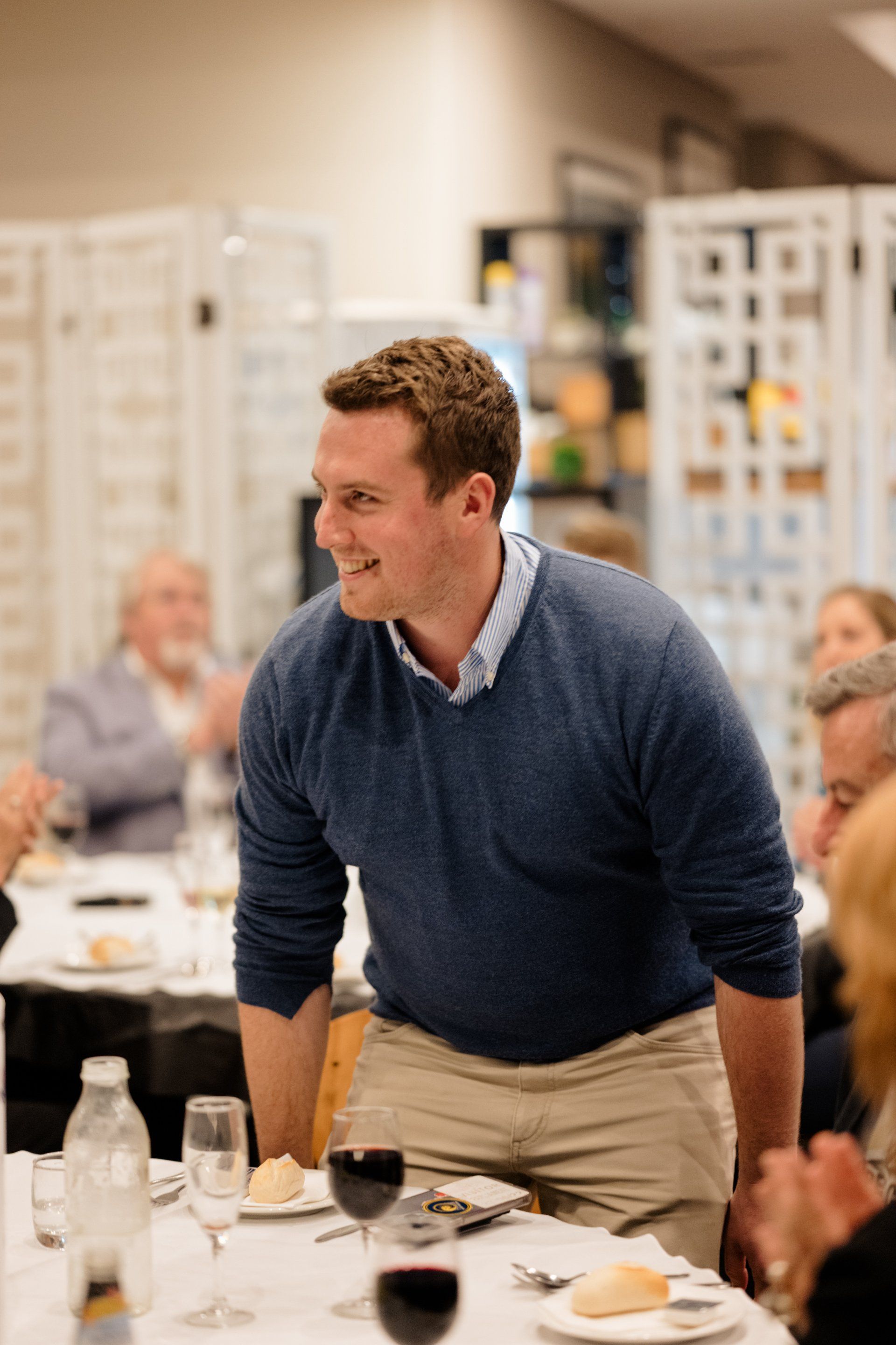 A man in a blue sweater is standing in front of a table with a glass of wine.