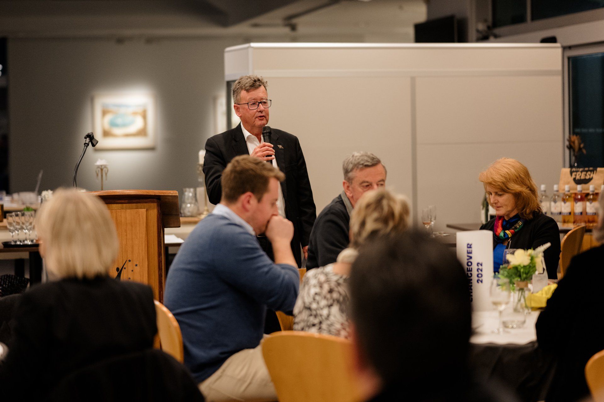 A man is standing at a podium giving a speech to a group of people sitting at tables.