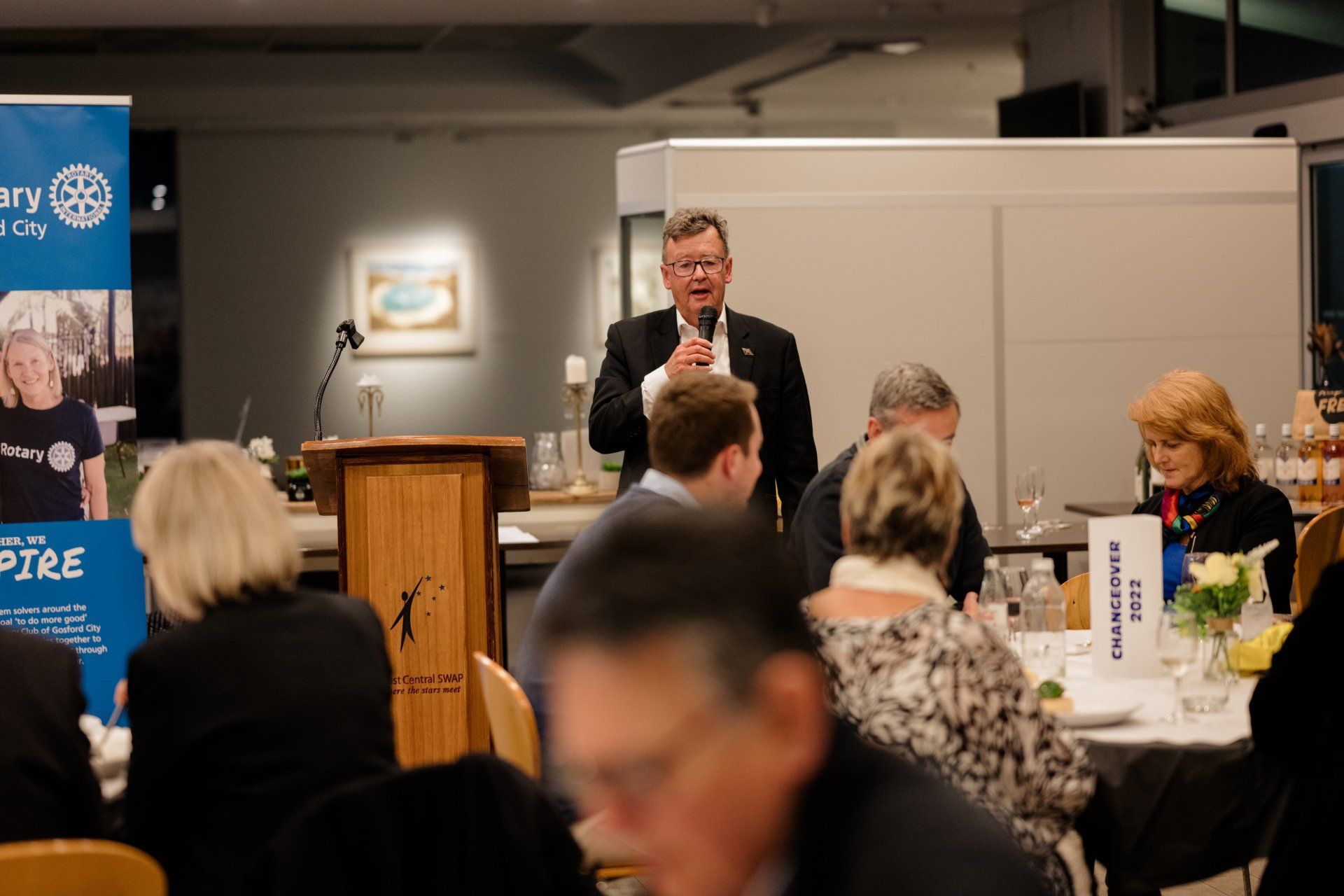 A man is standing at a podium giving a speech to a group of people sitting at tables.