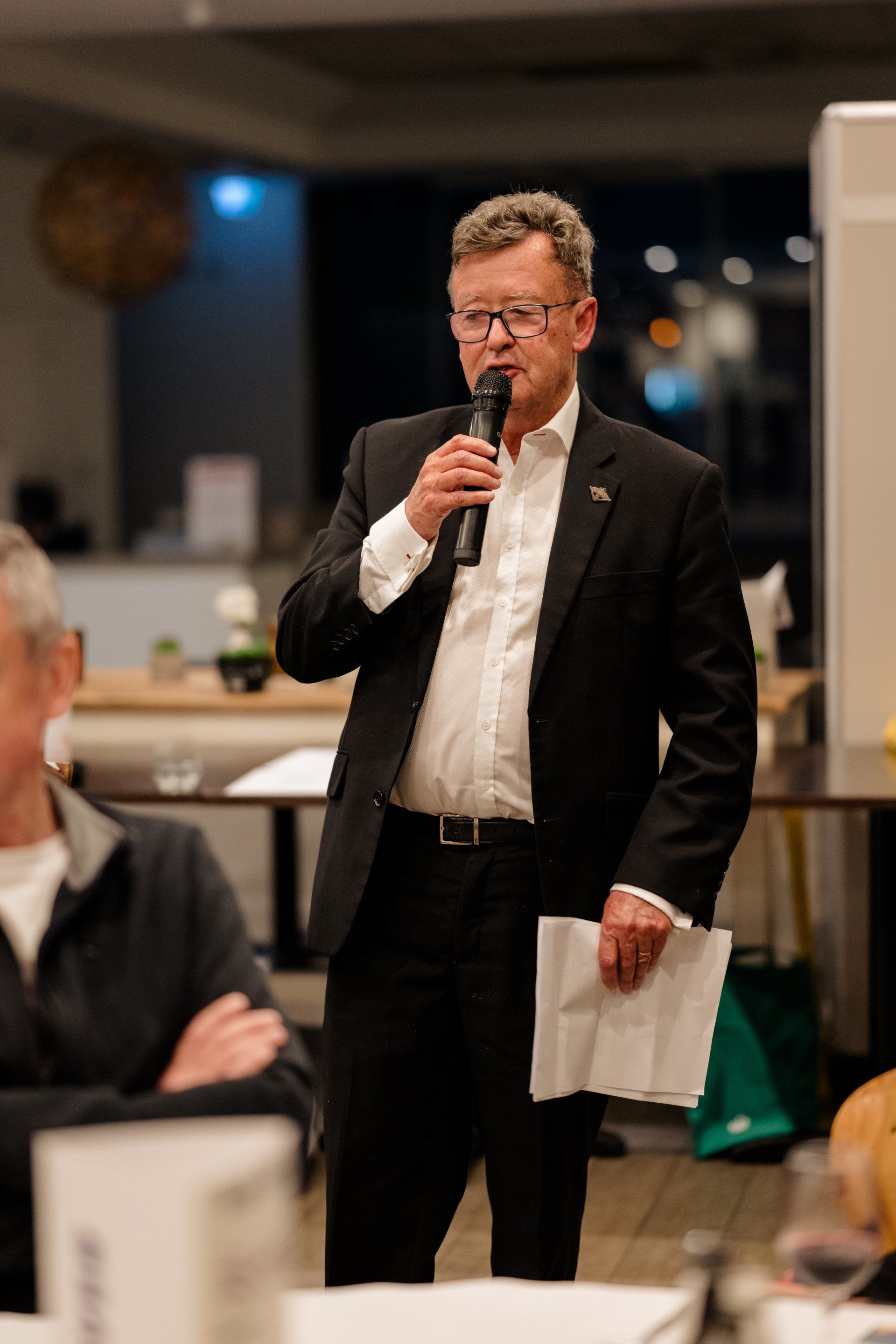 A man in a suit is speaking into a microphone while holding a piece of paper.