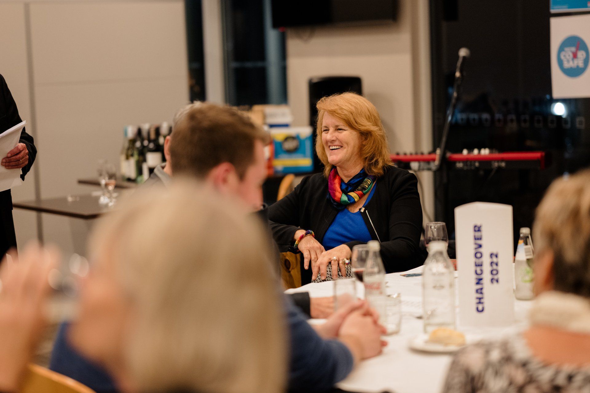 A woman is sitting at a table laughing while a man stands behind her.