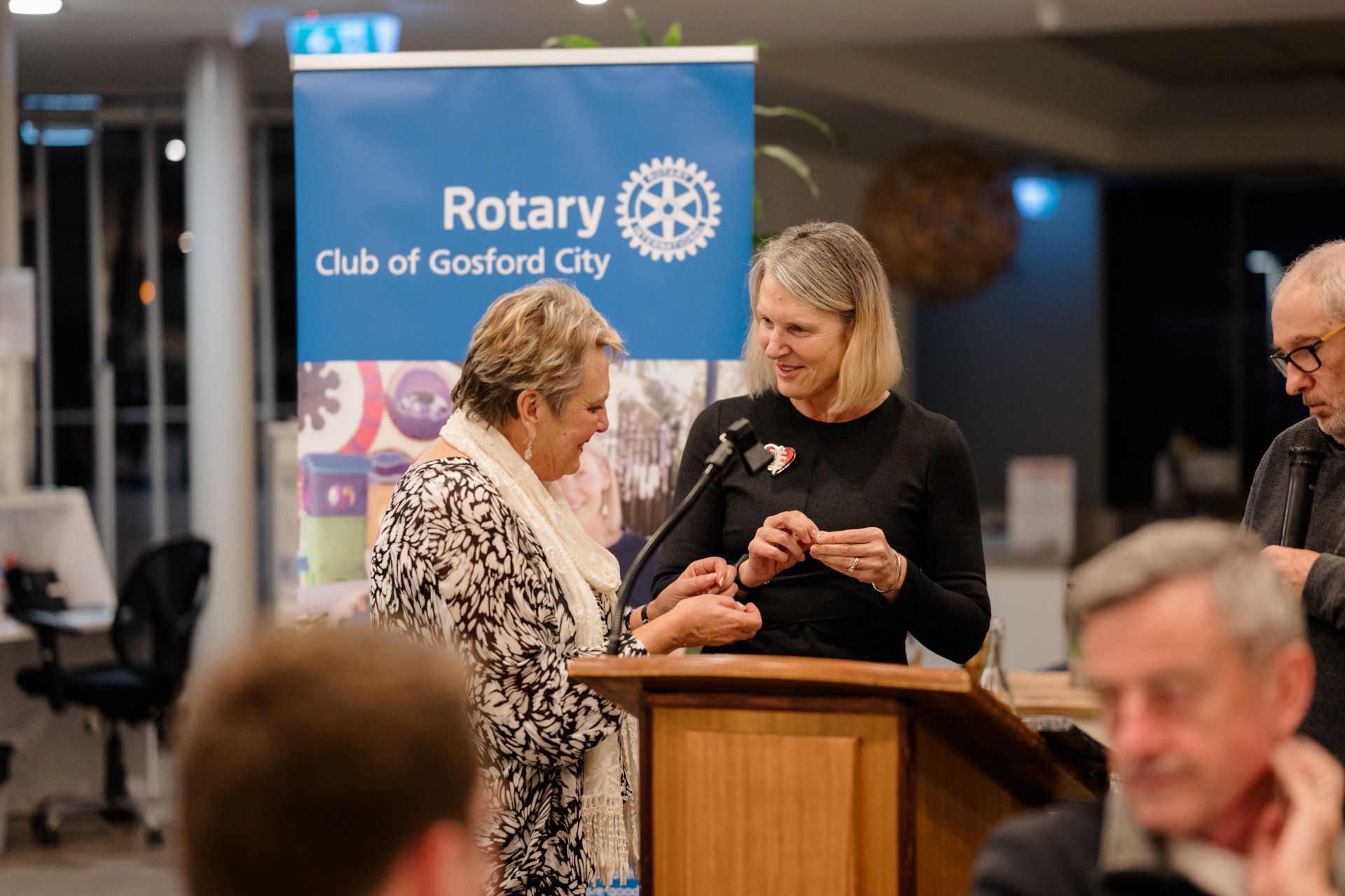 Two women are standing at a podium in front of a sign that says rotary club of gosford city.