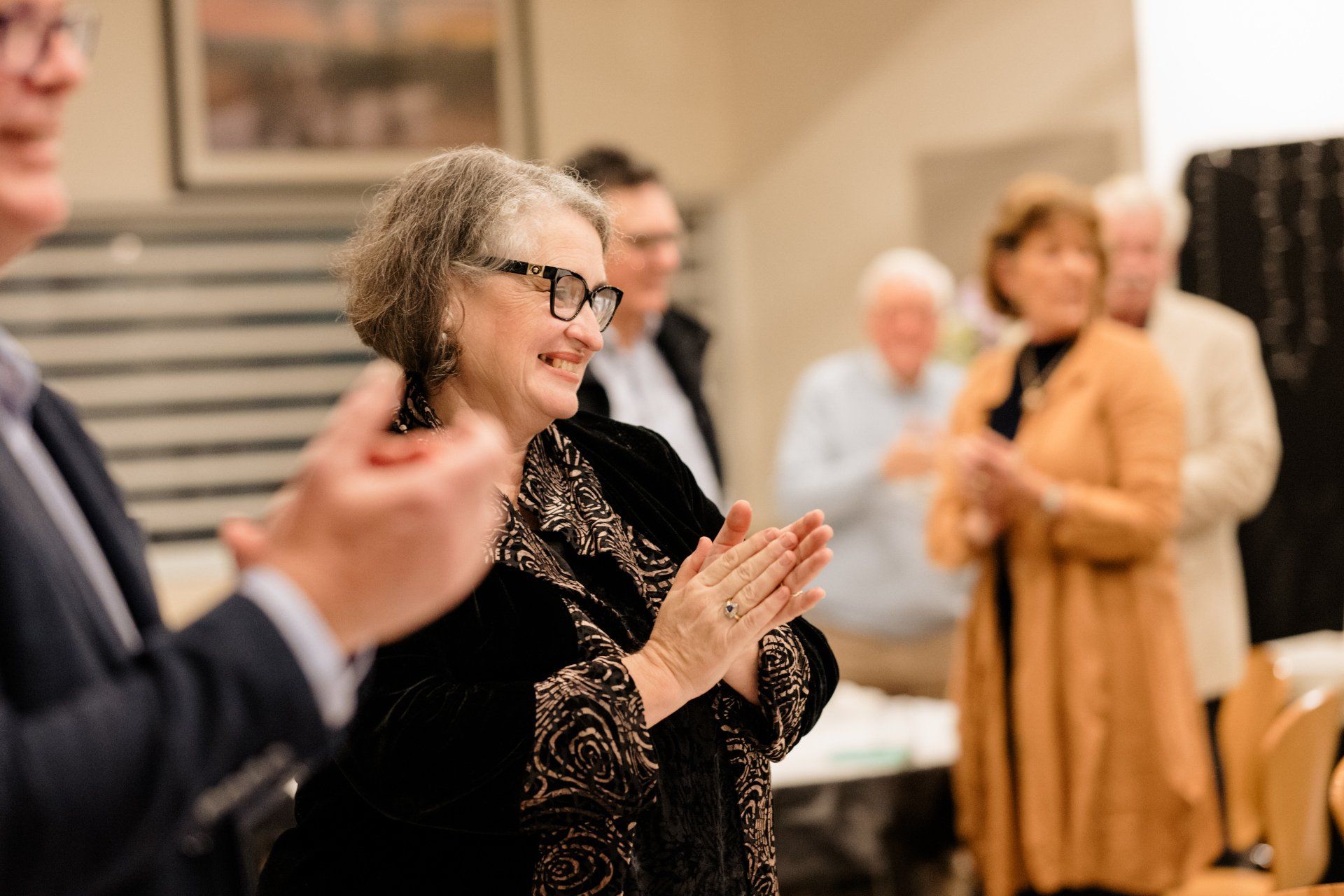 A woman wearing glasses is applauding while standing in front of a group of people.