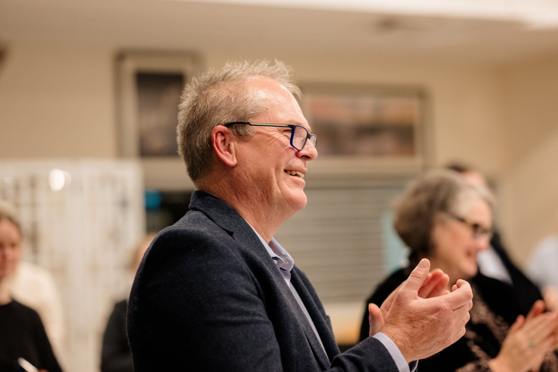 A man in a suit and glasses is clapping his hands in a room.