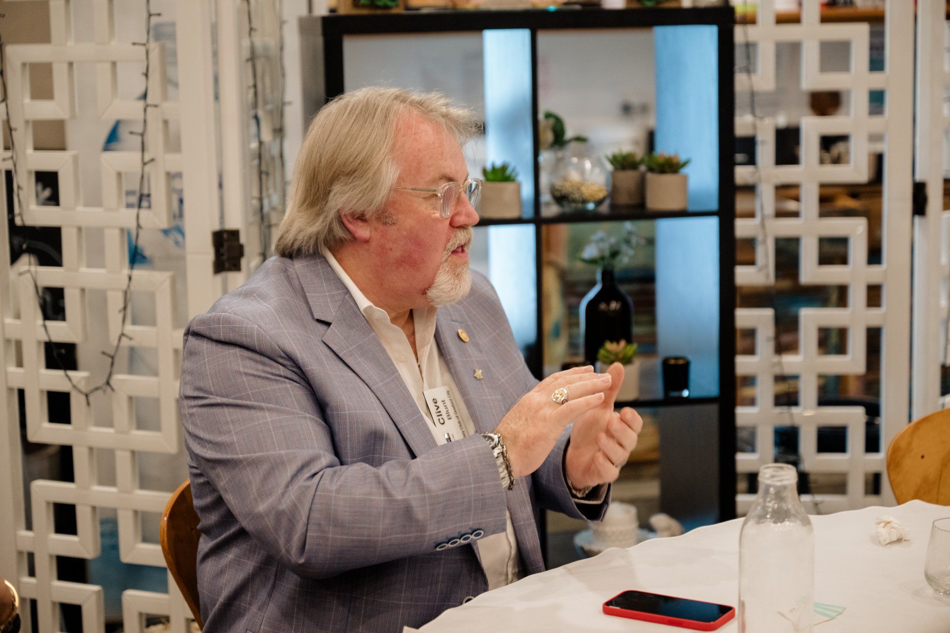A man in a suit and glasses is sitting at a table talking to someone.