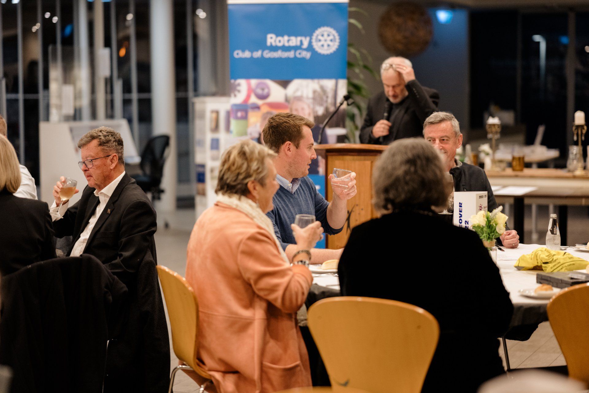 A group of people are sitting at tables in front of a rotary sign.