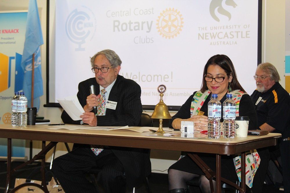 A group of people are sitting at a table in front of a screen that says central coast rotary clubs
