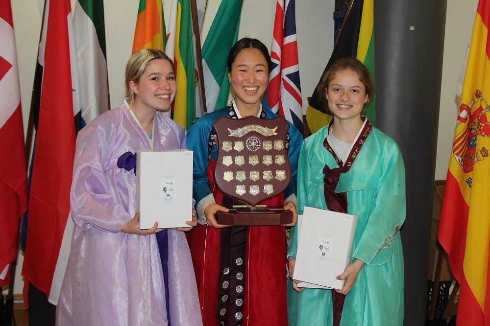 Three girls are holding a plaque in front of flags.