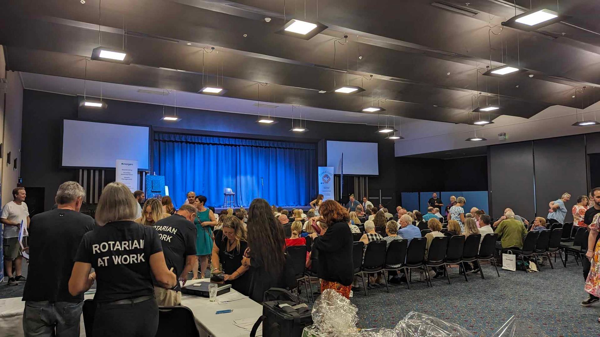 A large room filled with people sitting in chairs and standing in front of a stage.