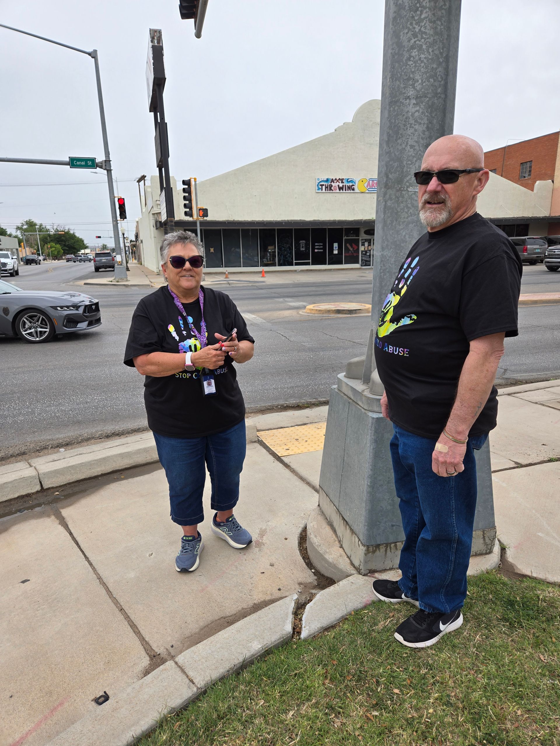 Two people standing on a sidewalk near a street corner, one beside a gray pole and one near the curb.